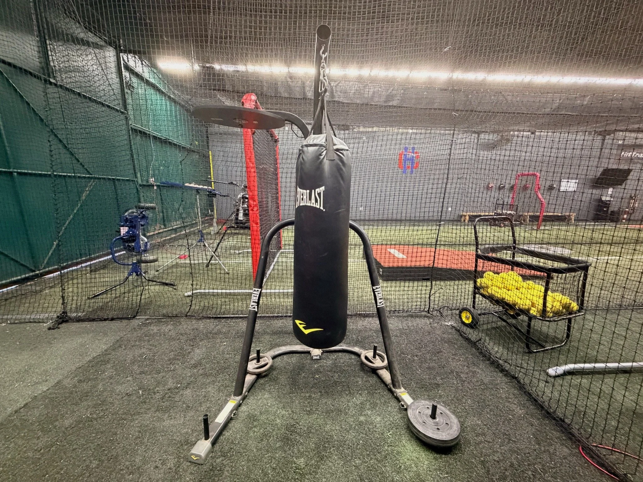 Indoor sports training area with a punching bag, weight sled, and various gym equipment behind a netted barrier.