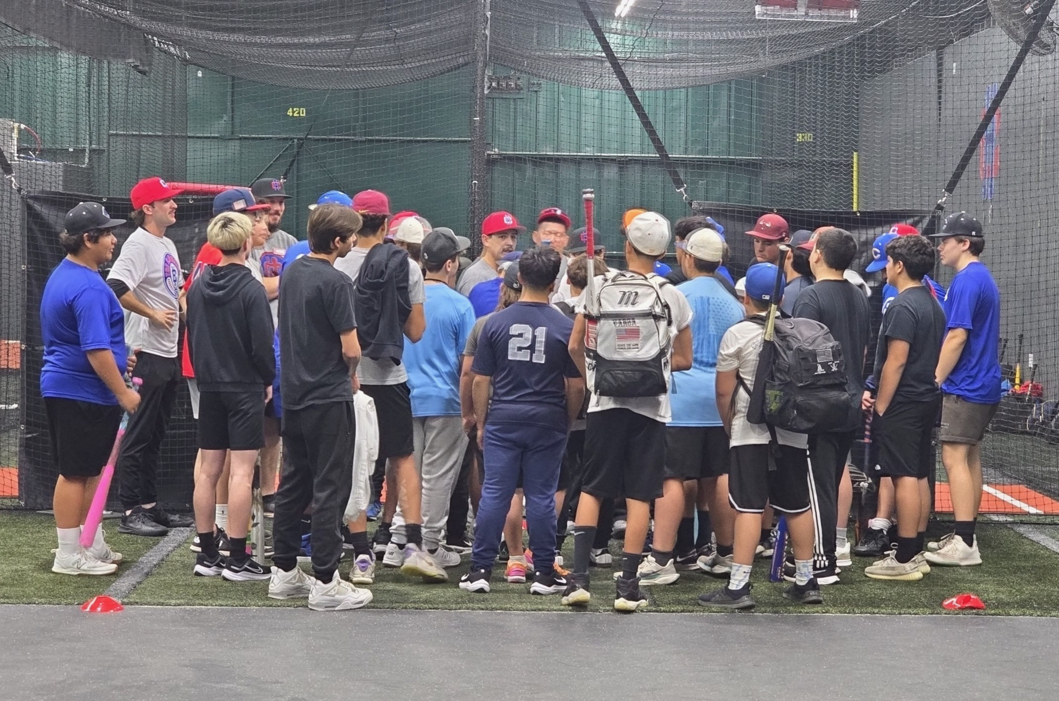 A large group of young baseball players and coaches gathered inside a batting cage, listening to instructions or a discussion, with some holding baseball bats and wearing different colored uniforms and gear.
