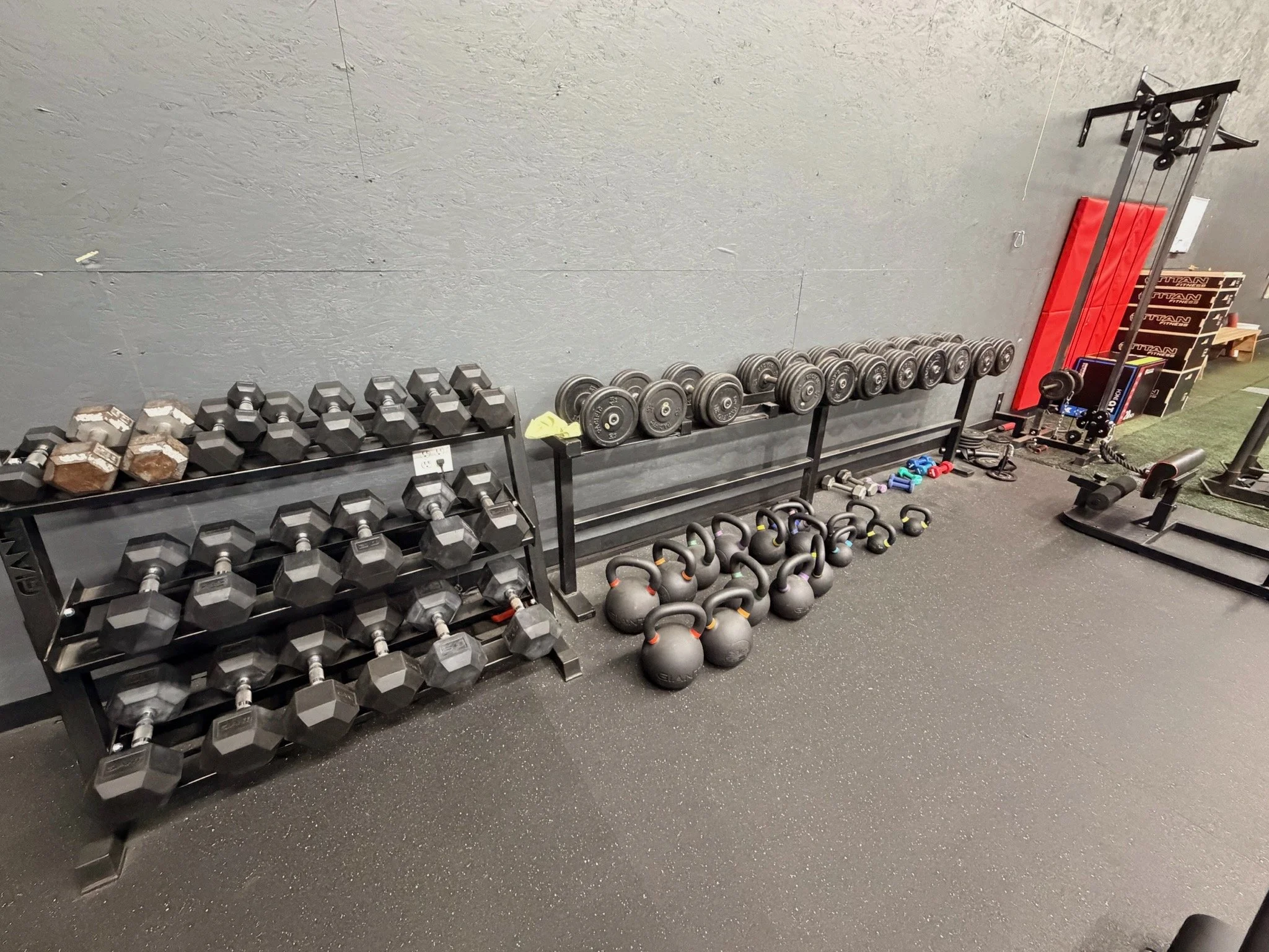 Gym equipment including dumbbells, kettlebells, weight plates, resistance bands, and workout mats arranged against a gray wall.