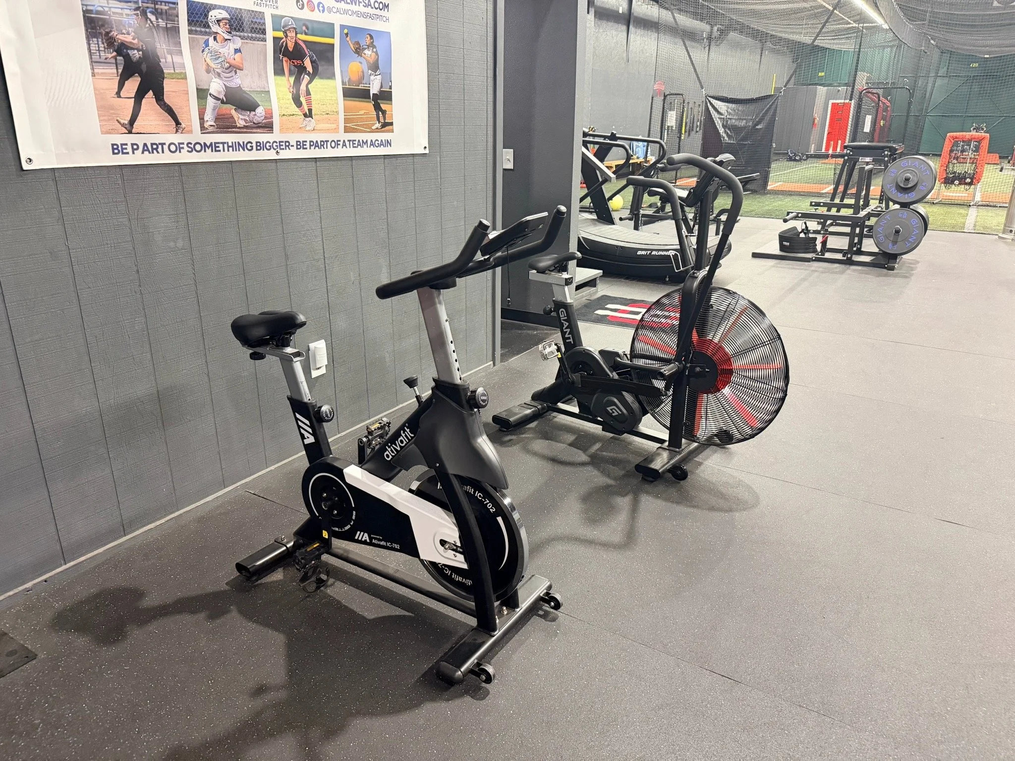Indoor gym with stationary exercise bike and elliptical trainer in front of a wall with a motivational poster, and a sports field visible through the glass exit door.
