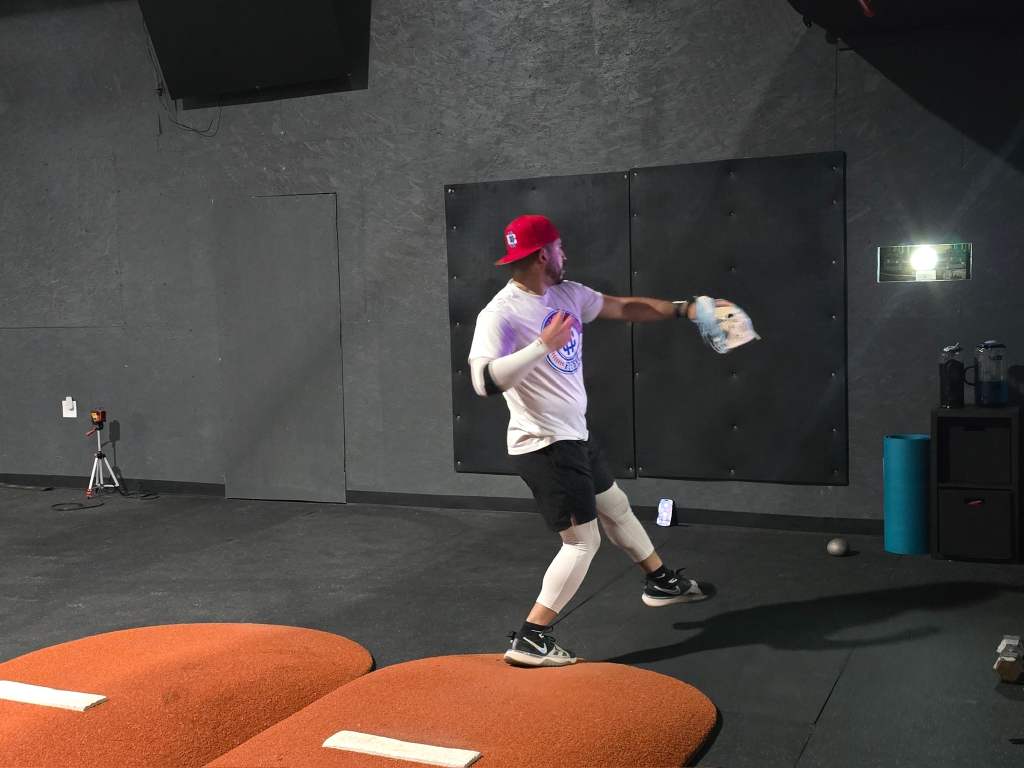 Man practicing baseball in gym, wearing a red cap, white shirt, and leggings, throwing a baseball.