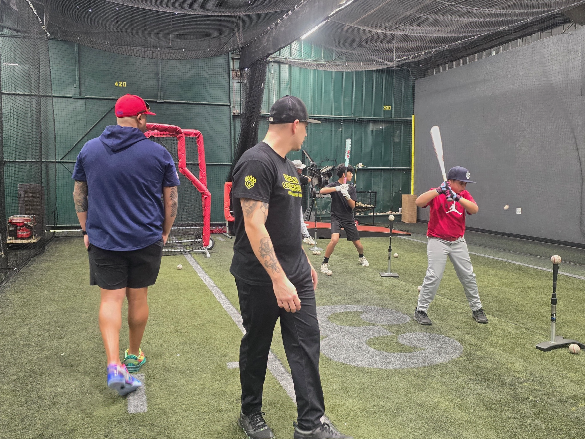 Group of young men practicing baseball swings in an indoor batting cage with artificial turf, pitching machines, and baseballs.