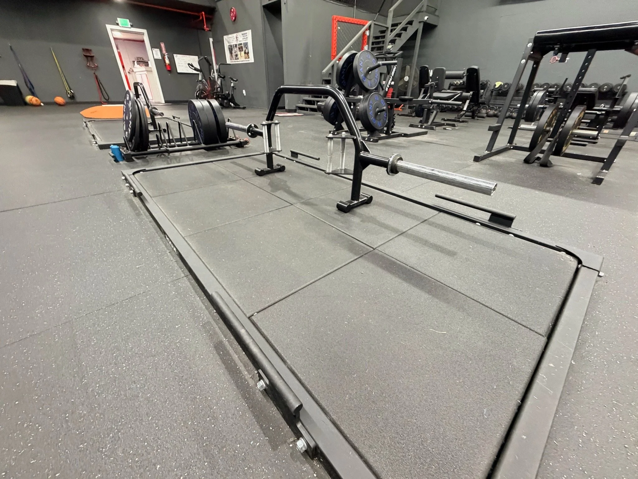 A gym with various weightlifting equipment, including a barbell with weight plates on a squat rack, weight plates stored on racks, and other exercise machines in the background.