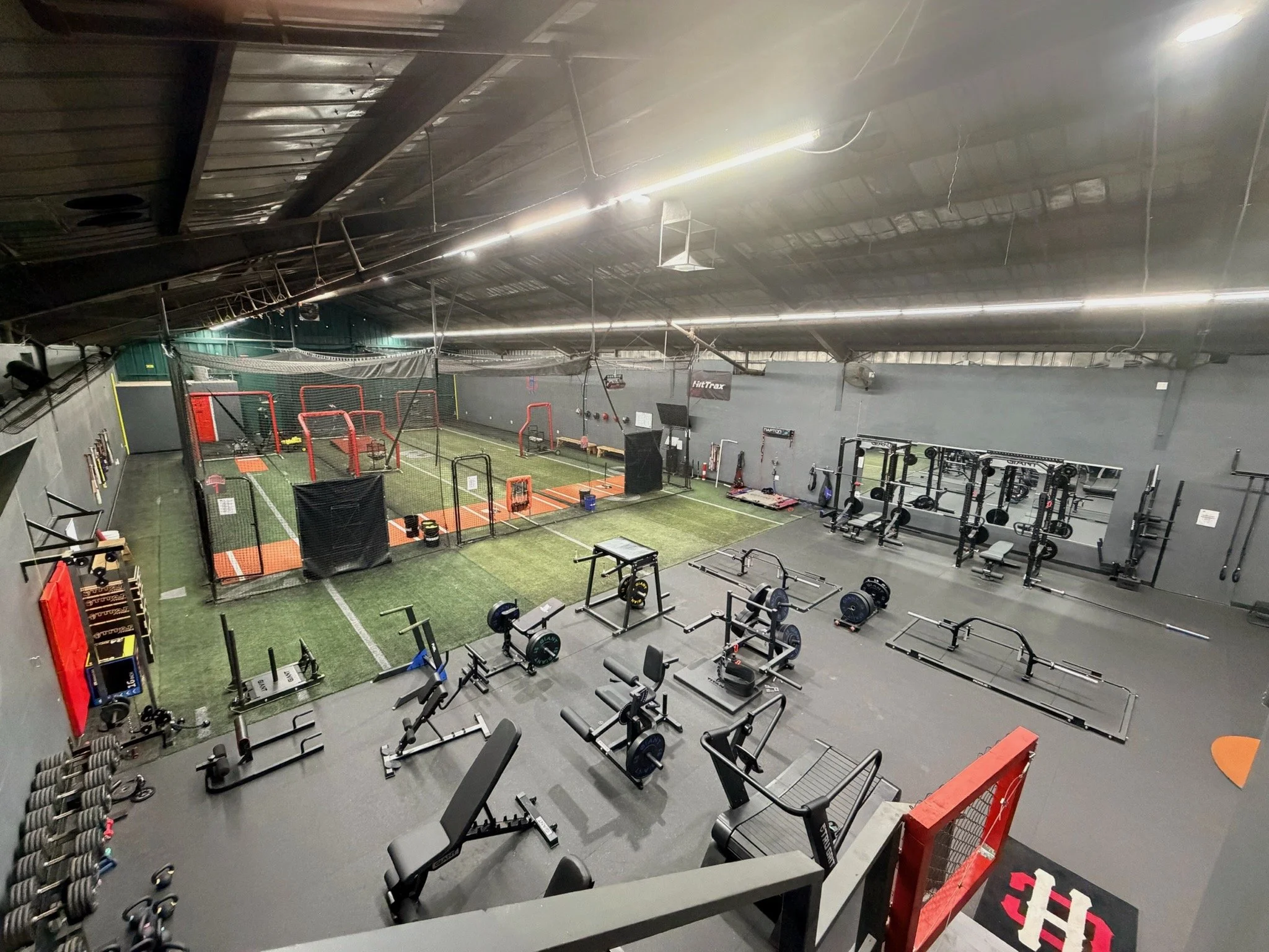 Indoor gym with weightlifting equipment and a turf area for baseball or softball training, including cages, racks, and benches.