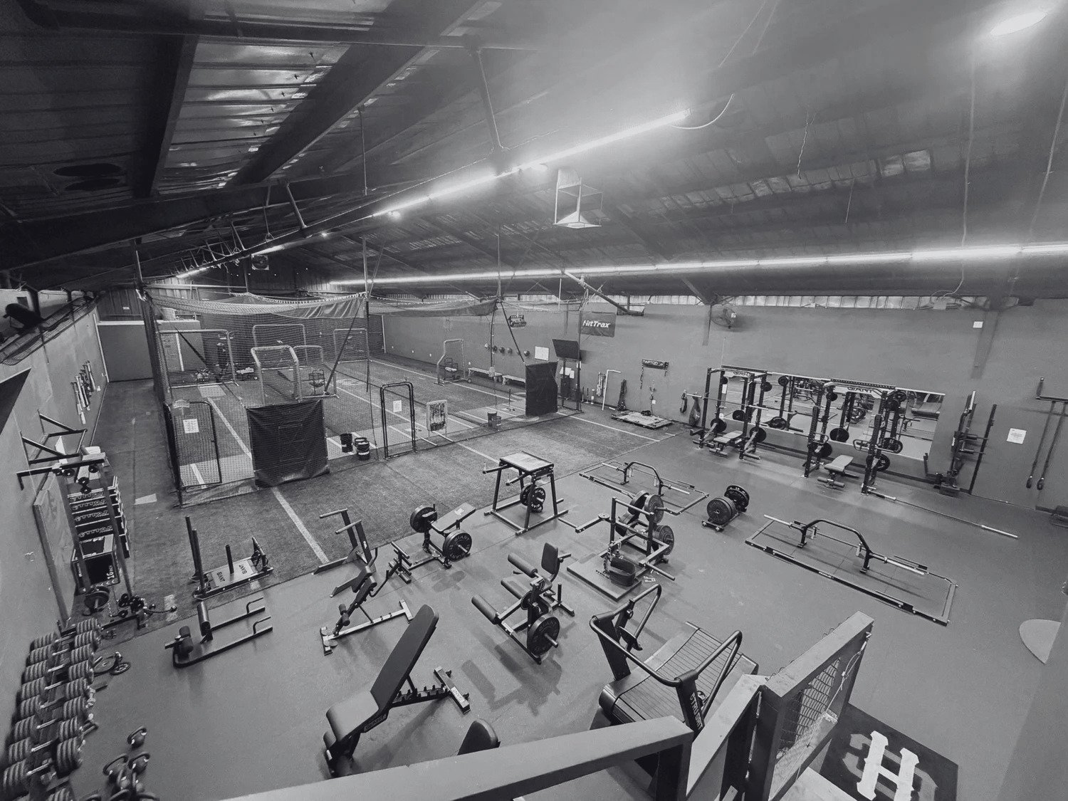 Empty indoor gym with weightlifting equipment, benches, barbells, and a fenced area for sports or training, lit by overhead fluorescent lights.