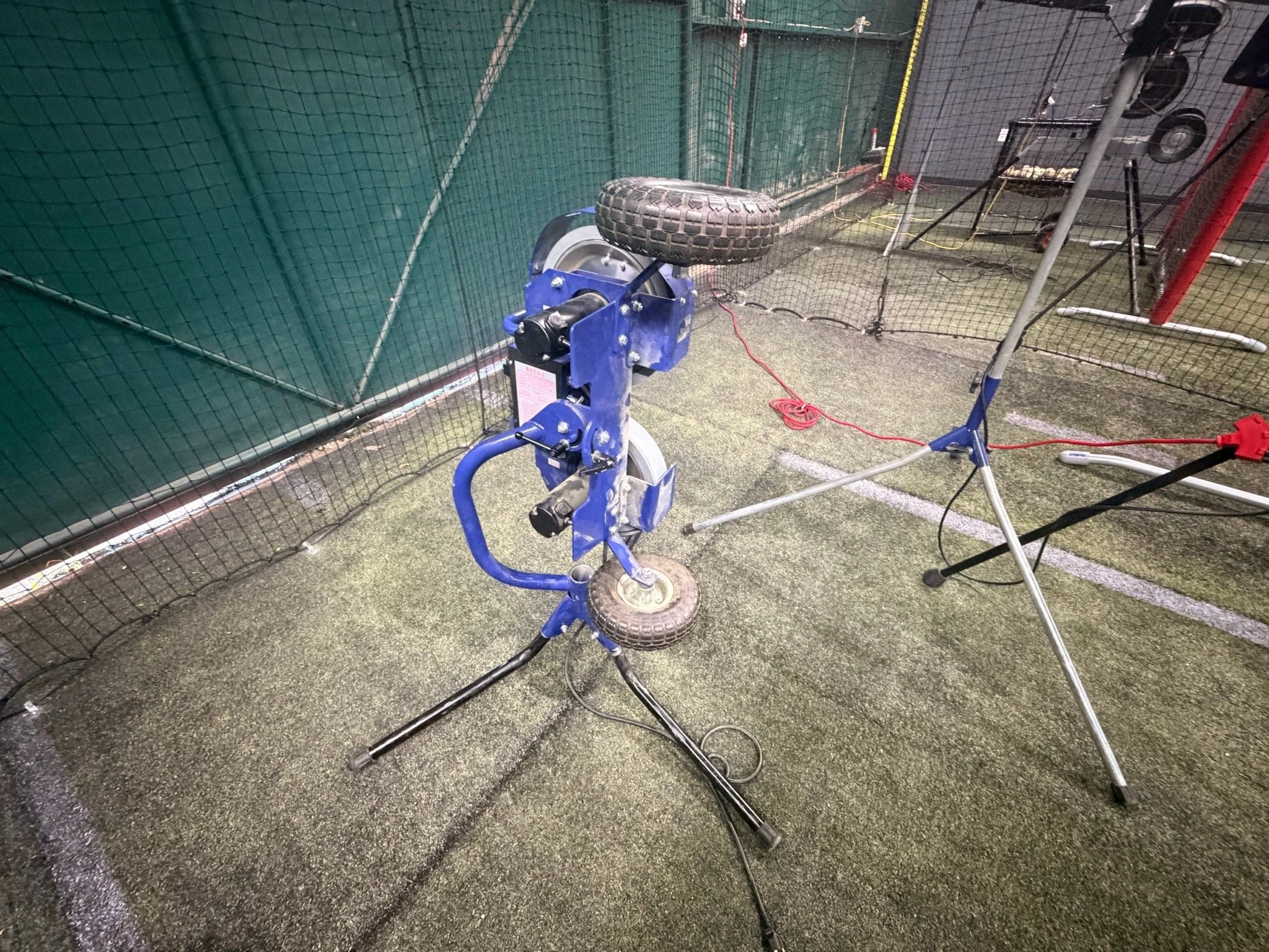 A blue pitching machine with a tire on top, on a wet indoor sports court, protected by a green netting.