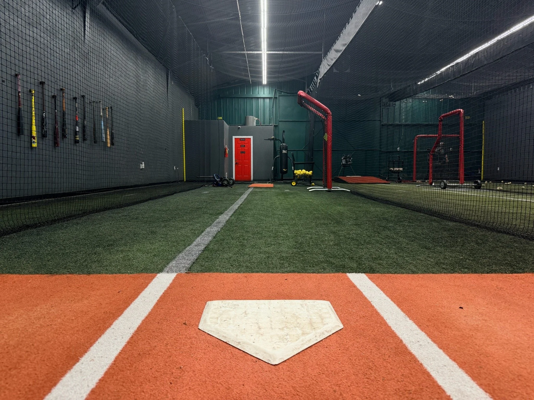 Indoor sports training facility with turf, pitching mound, netting, and training equipment such as baseballs, bats, and a pitching screen.