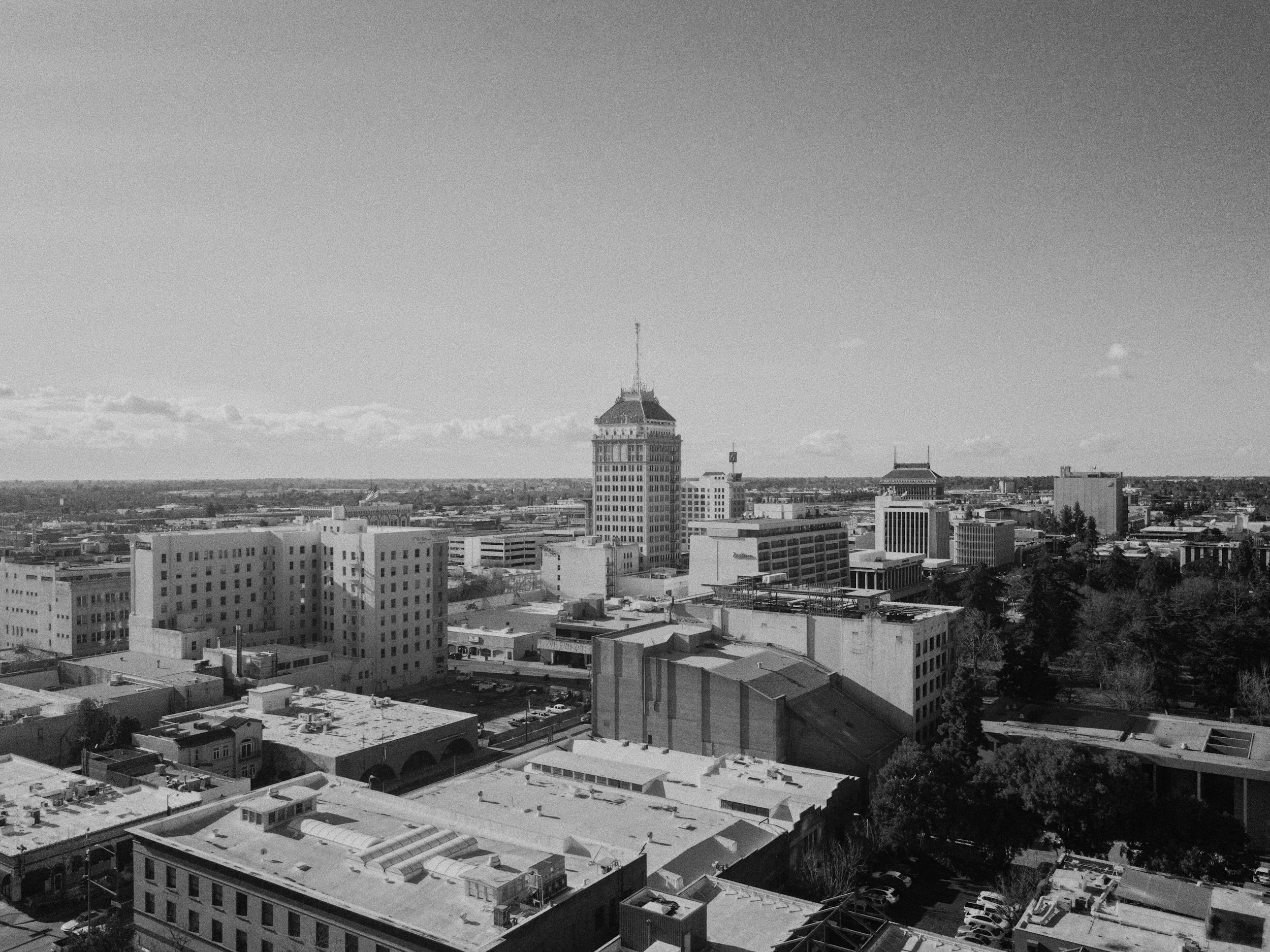 Black and white aerial view of a city skyline with tall buildings, a prominent tower with a flag, and a mix of smaller buildings and trees in the foreground and background.