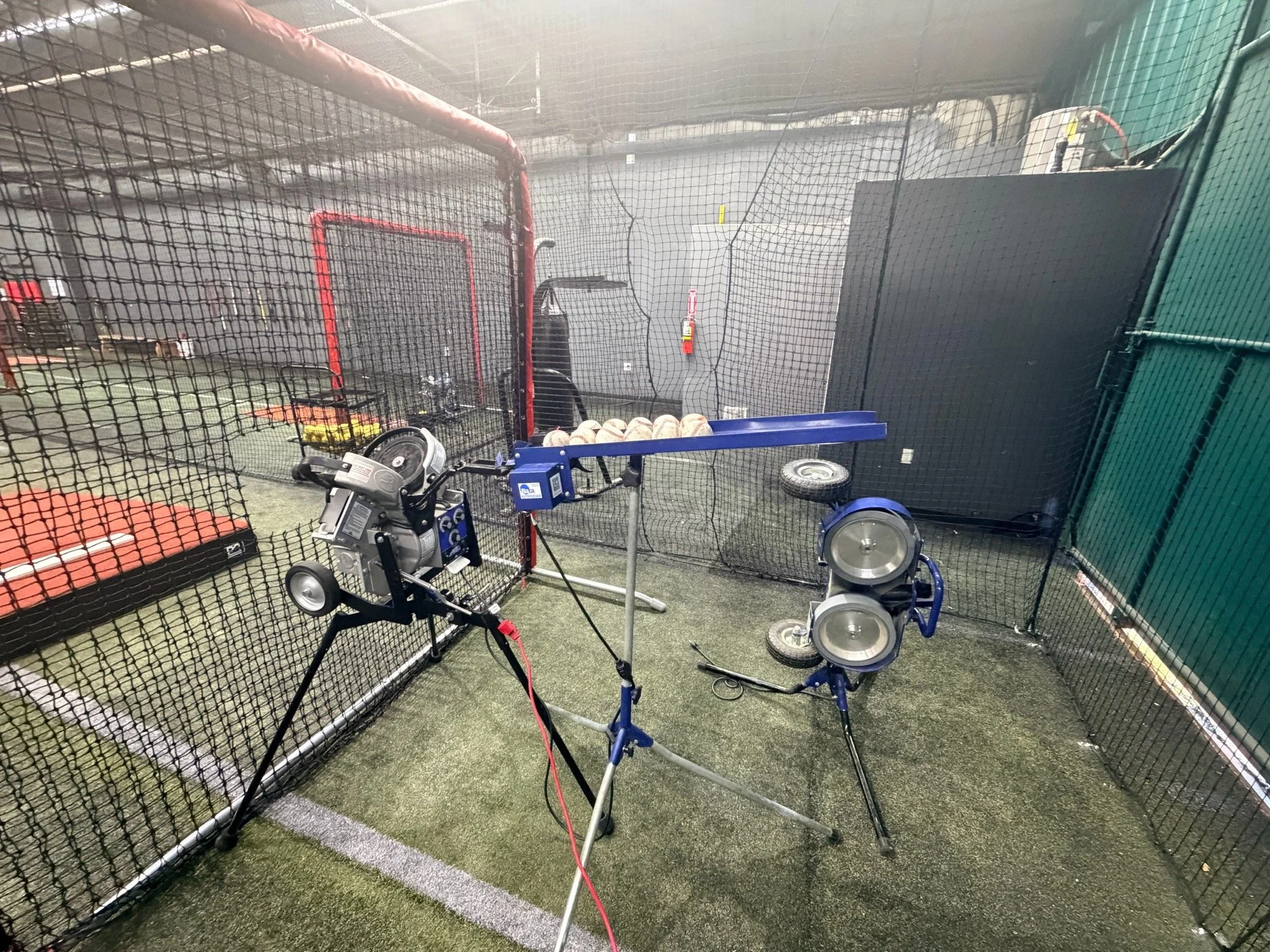 Indoor baseball or softball facility with pitching machine, baseballs on blue rack, and protective netting.