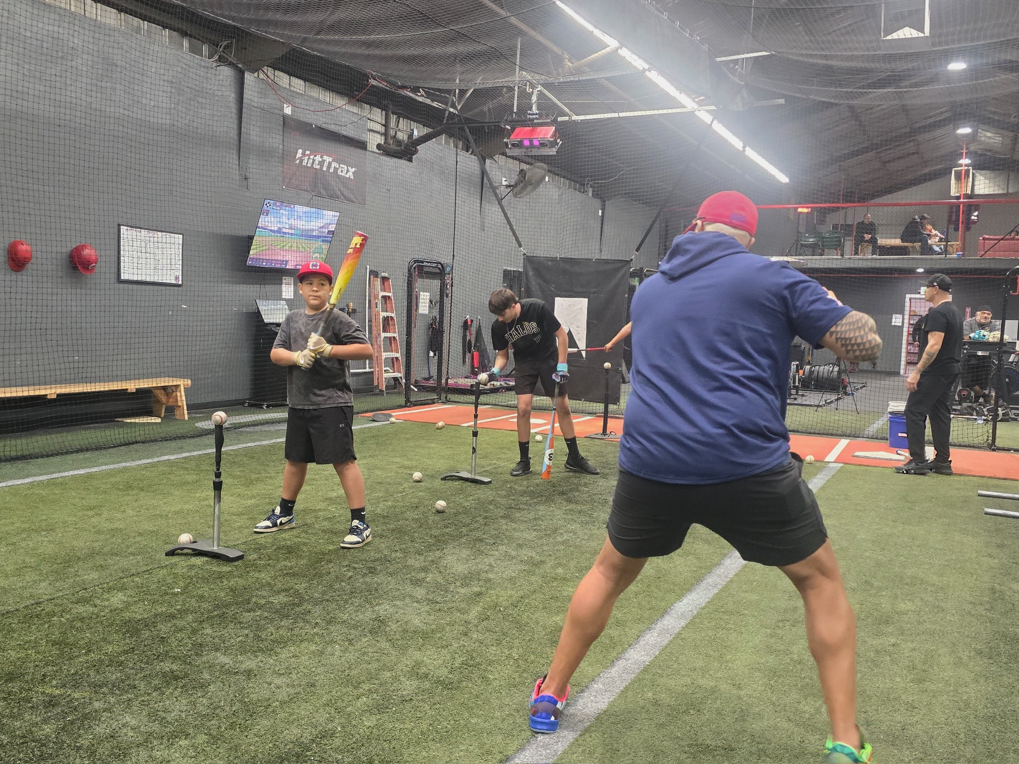 Children practicing baseball swings indoors with coach and batters, equipment, and batting practice stations.