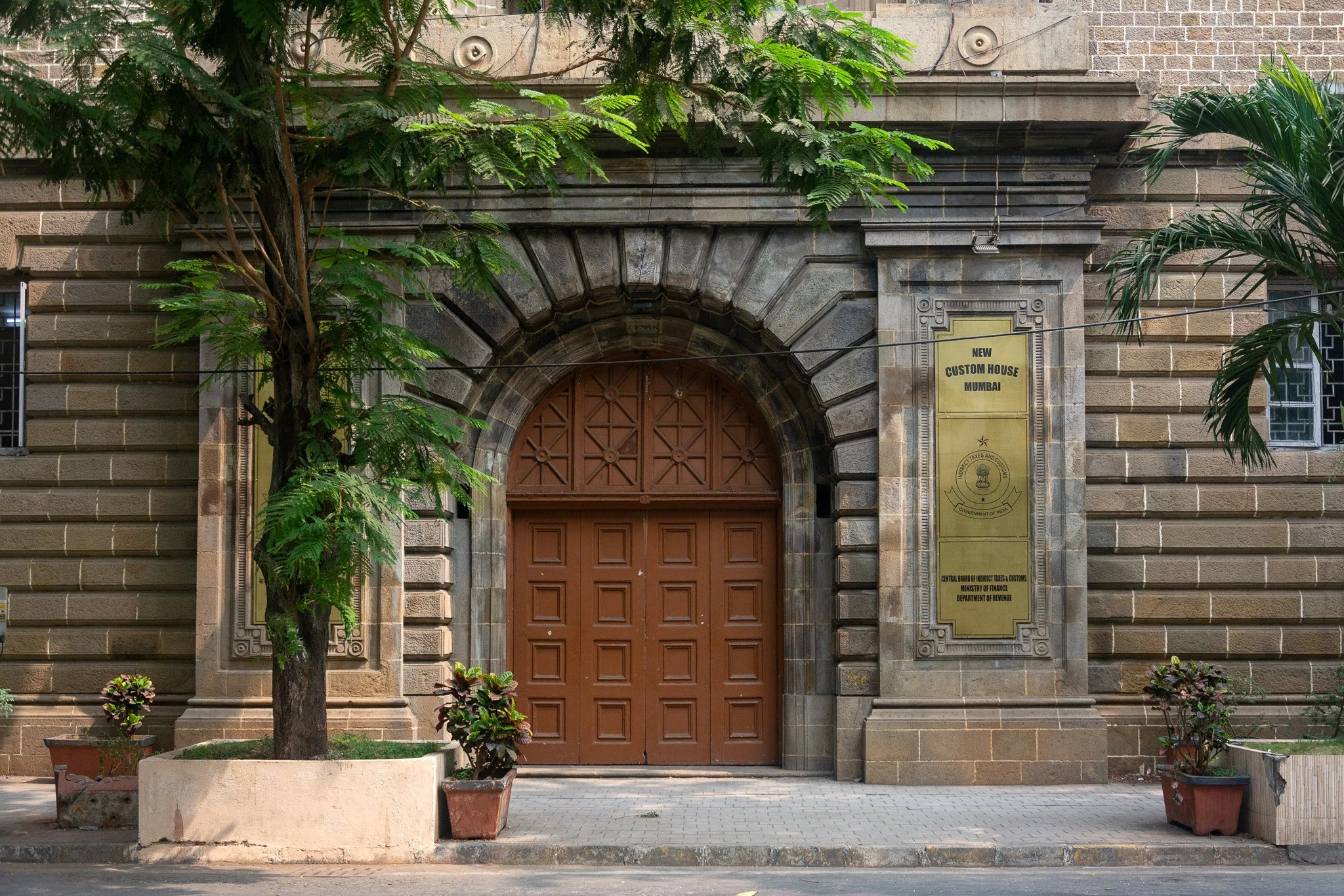 Brick building with stone arch and wooden door, a yellow sign reading "New Custom House Mumbai," and potted plants outside.