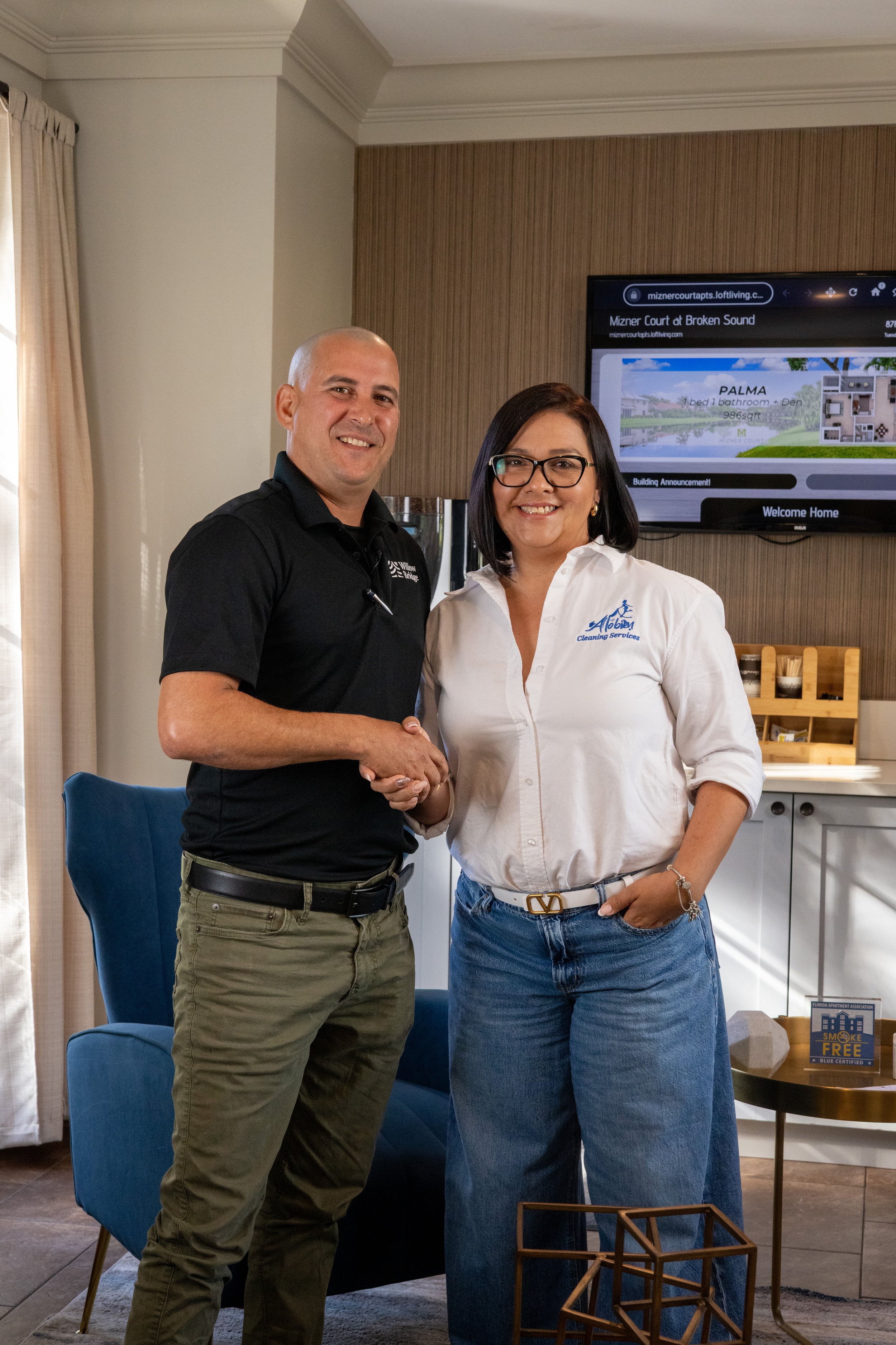 Man and woman shaking hands indoors, smiling, with a television screen behind them displaying a real estate website.
