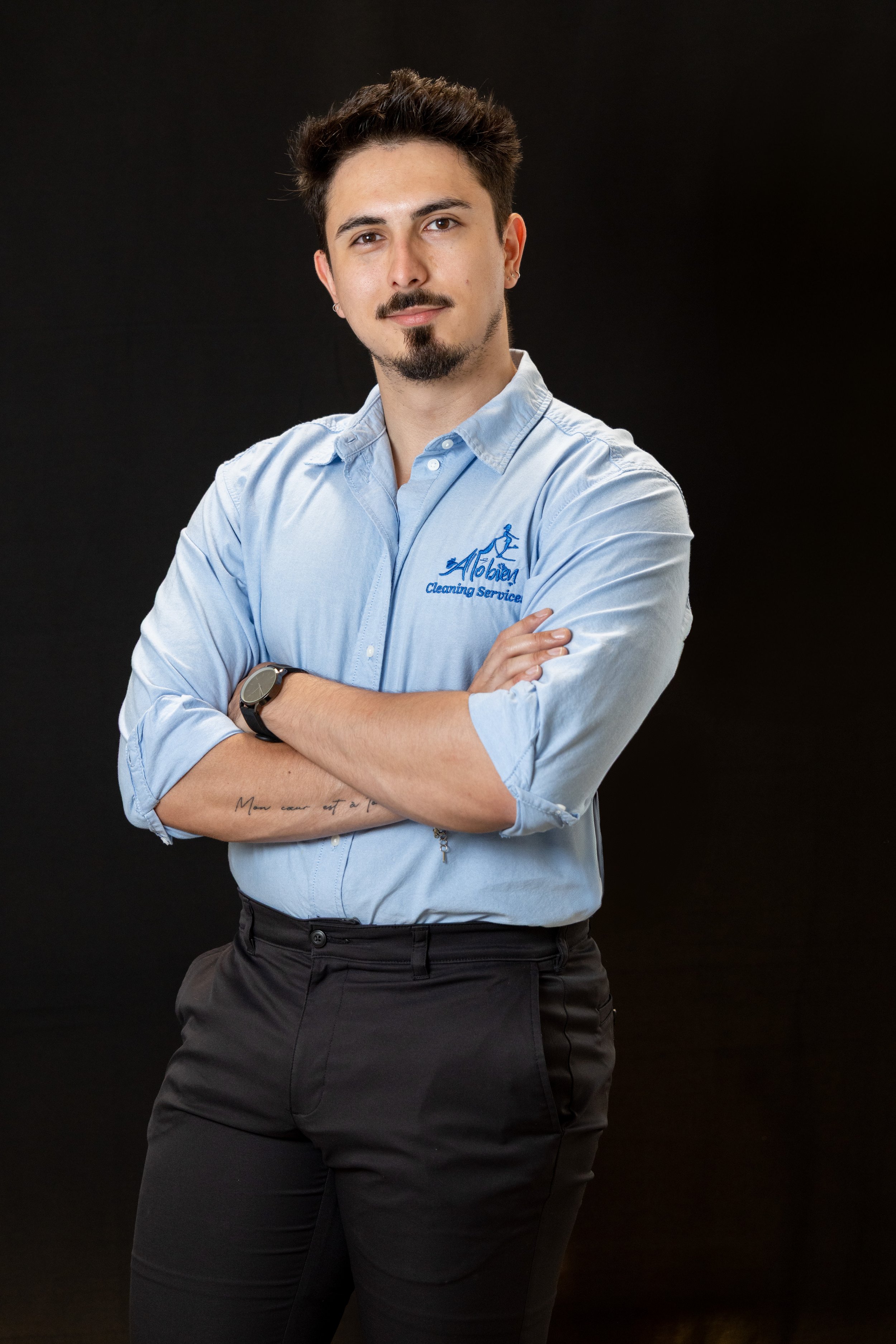 A man with dark hair and a beard, wearing a light blue shirt with a logo that says 'Alobien' and 'Cleaning Service', stands with arms crossed against a black background.