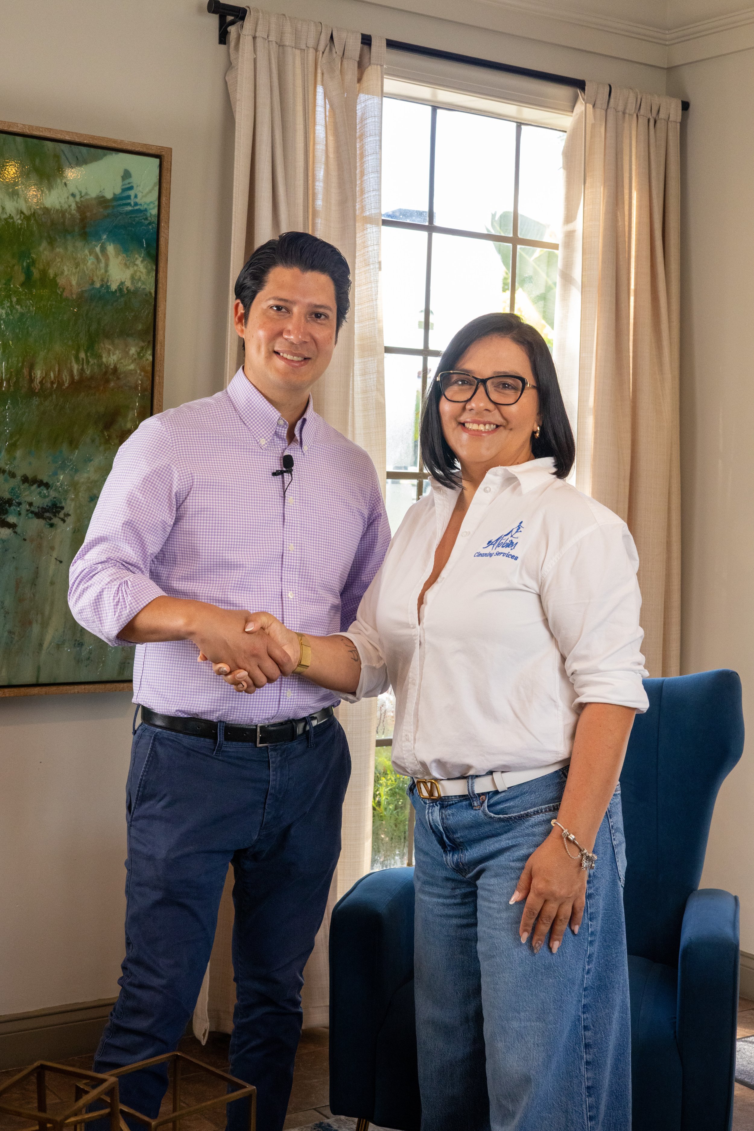 A man and woman shaking hands indoors, smiling, with a window and a painting in the background.