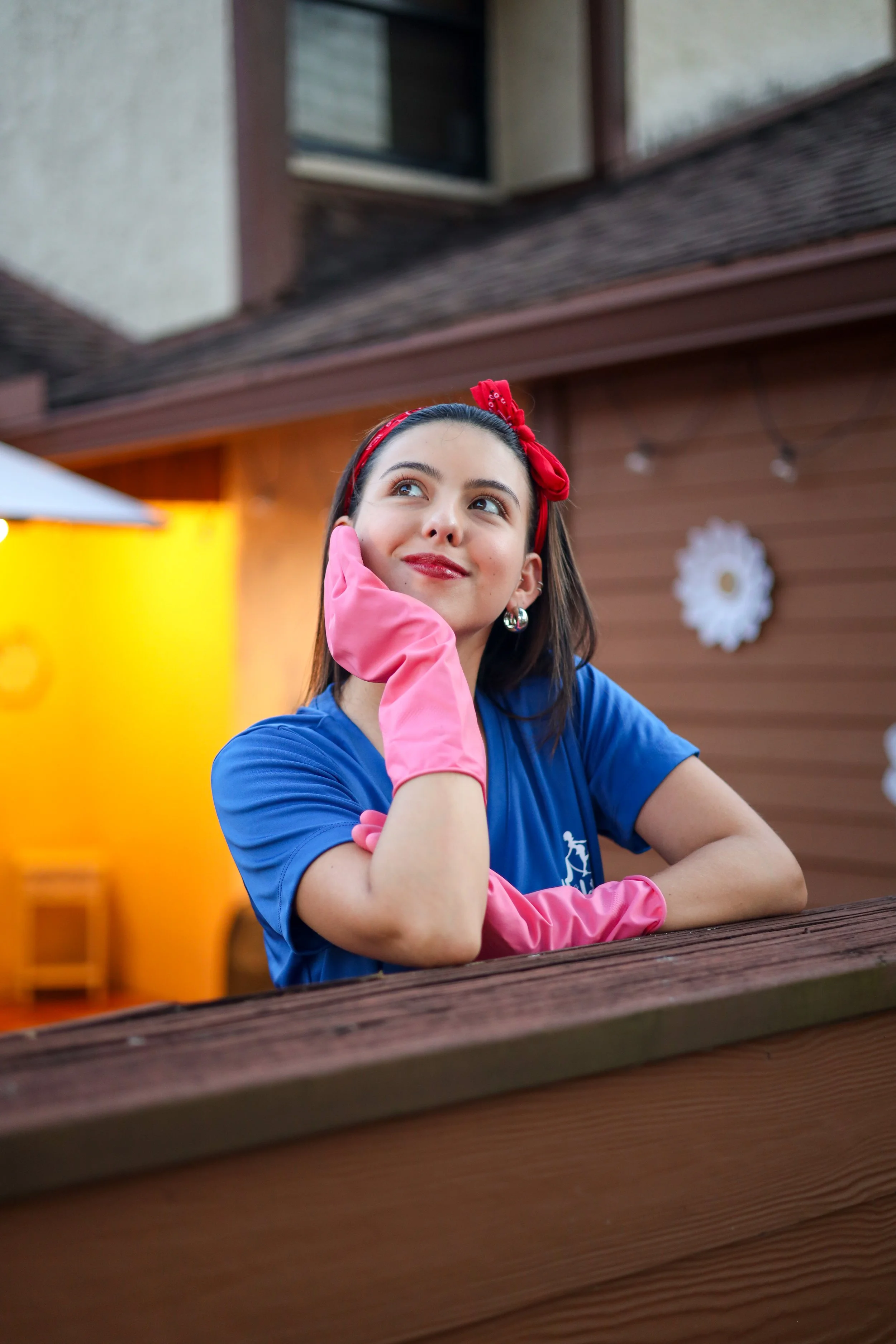 A young woman with long dark hair wearing a blue shirt, pink rubber gloves, and a red headband with a bow, sitting outdoors at a wooden table with a thoughtful expression.
