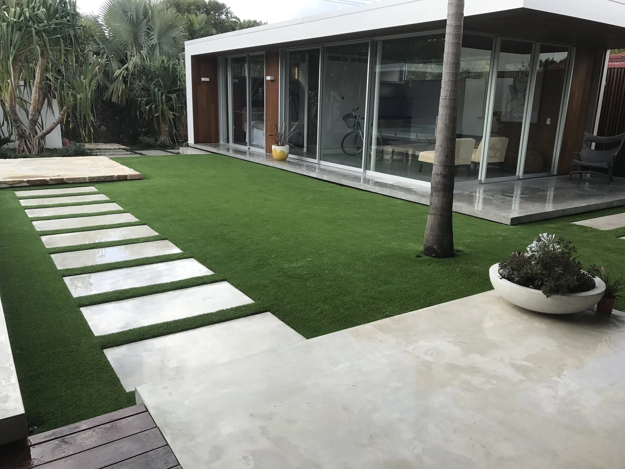 Modern backyard with a synthetic grass lawn, stone pathway, tropical plants, and a house with glass sliding doors and a patio.