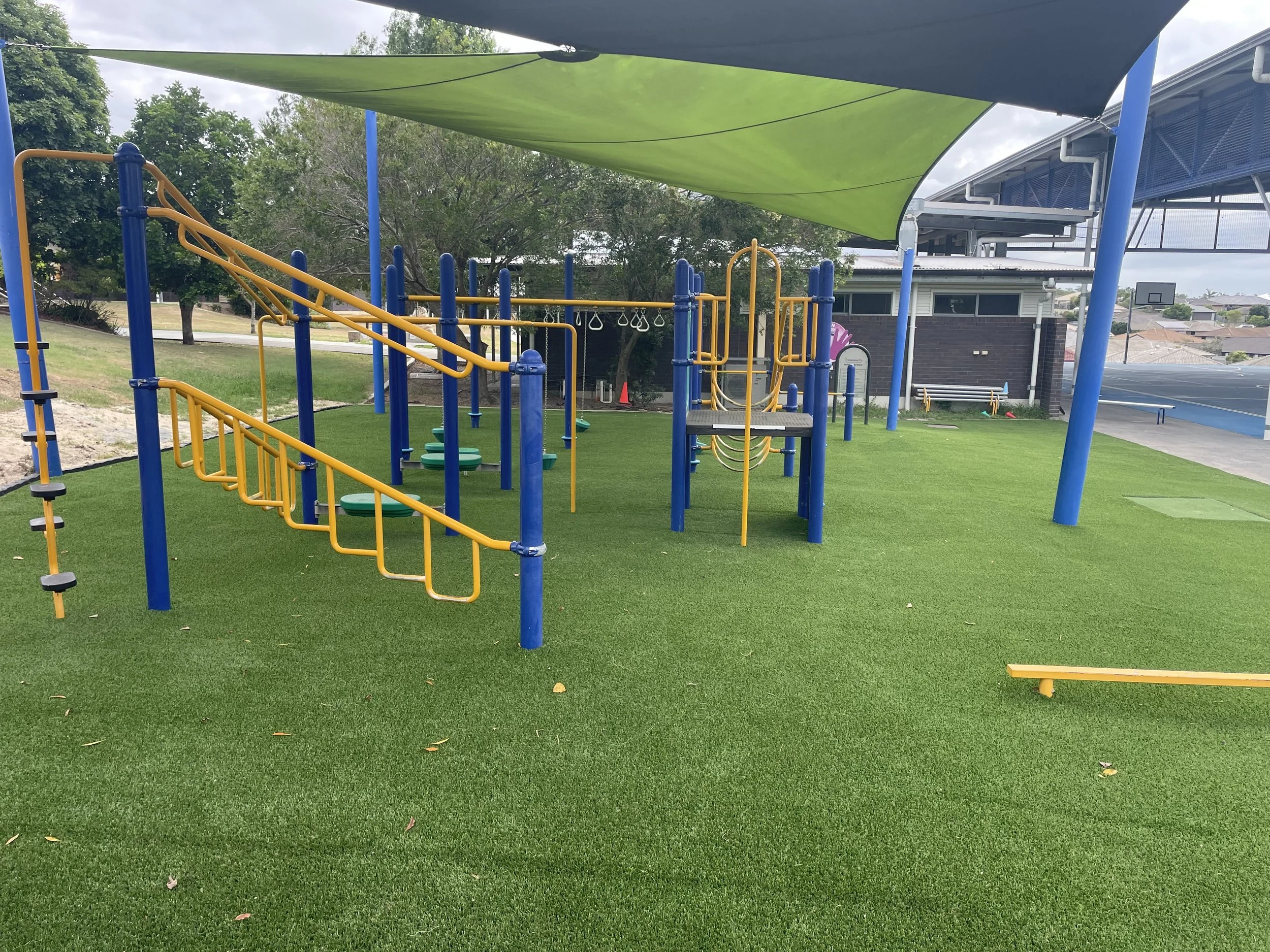 School playground with synthetic grass, green shade cloth, and blue and yellow play structures, near a basketball court and building.
