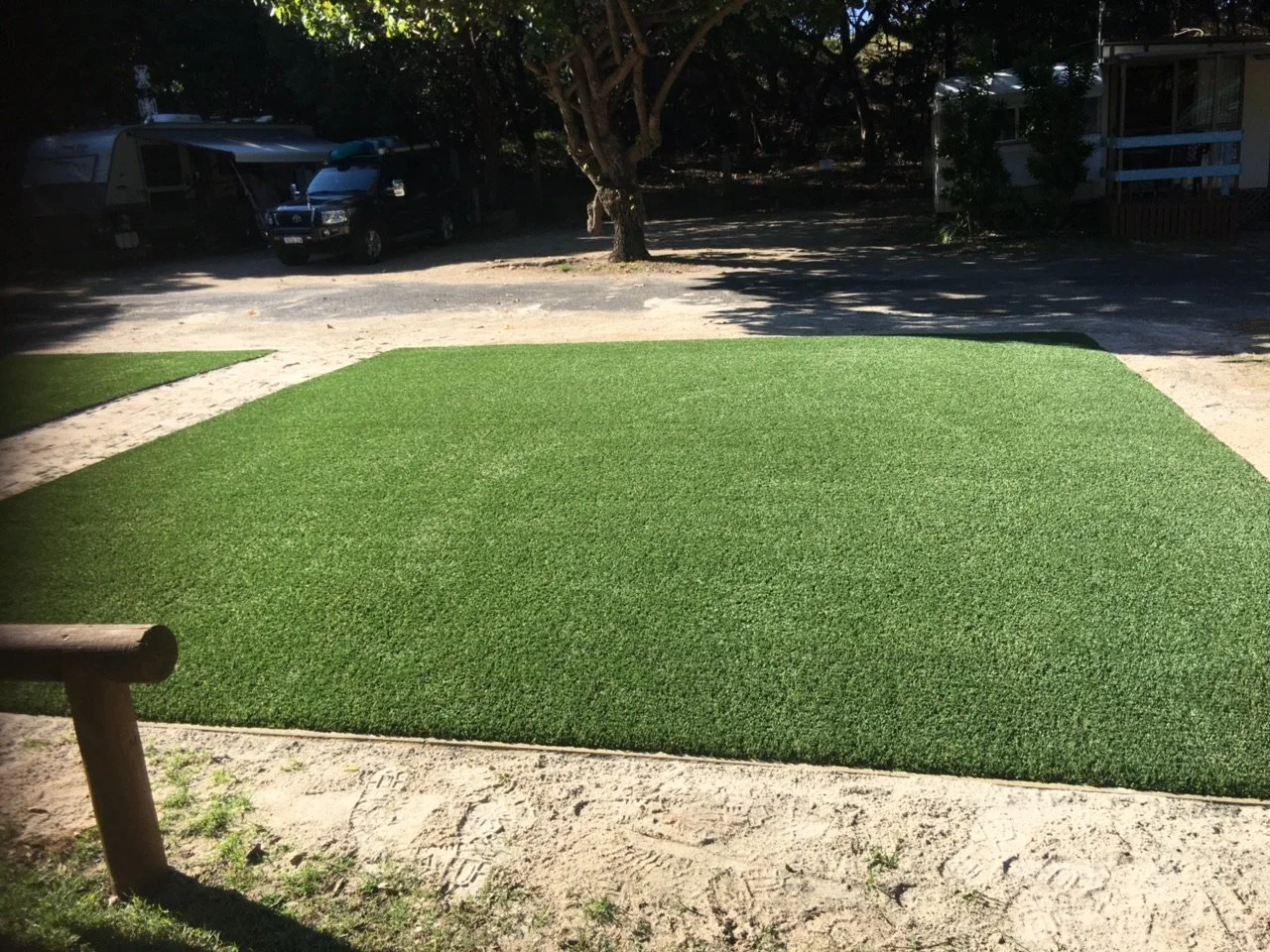 Synthetic green grass patch on sandy ground in outdoor setting, with trees, vehicles, and a building in the background.