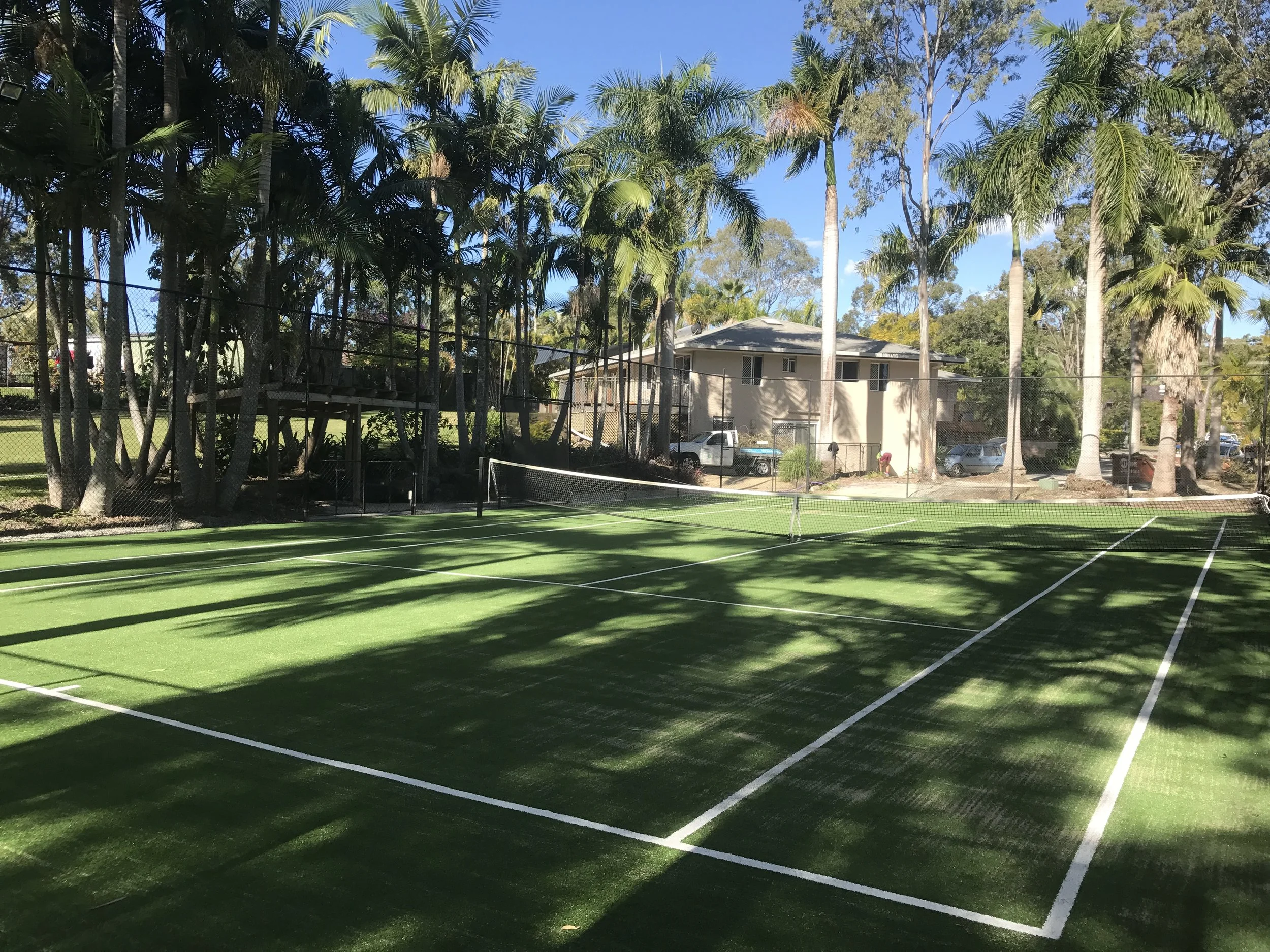 Synthetic grass tennis court with green surface, surrounded by tall palm trees and a fence, with a residential building and parked cars in the background.