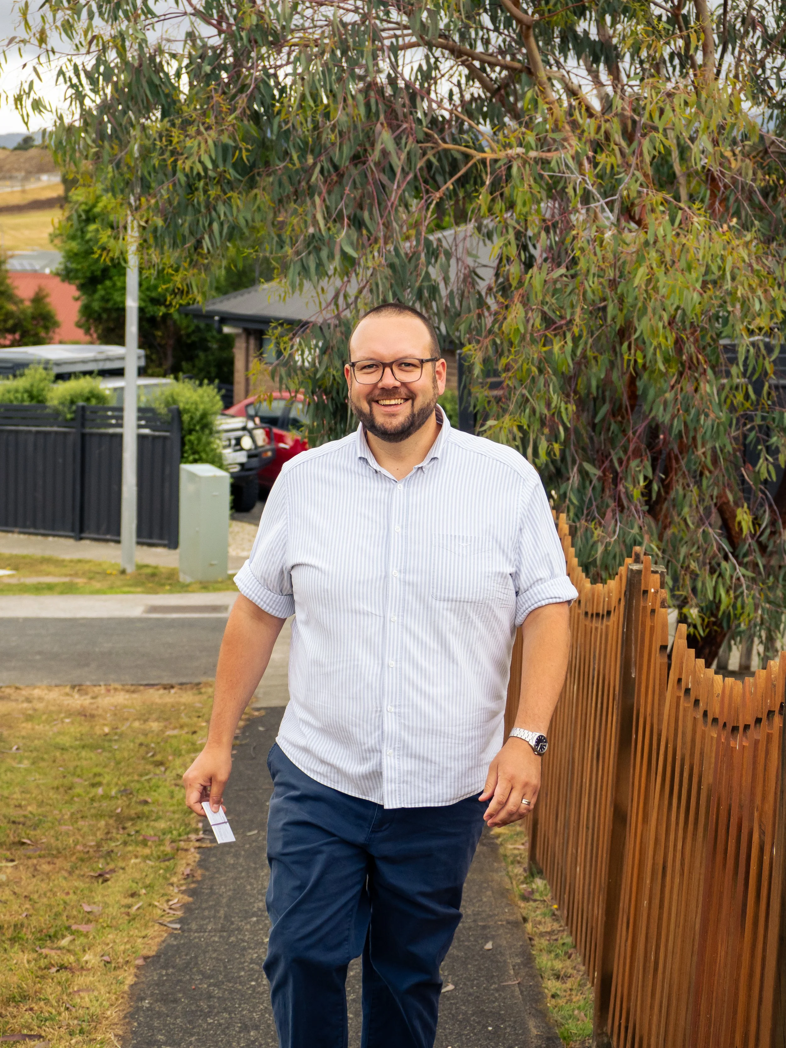 Tyler Petersen, wearing glasses, and with a short beard, walking on a sidewalk in the Tasmanian division of Huon alongside a wooden fence, with trees and parked cars in the background.