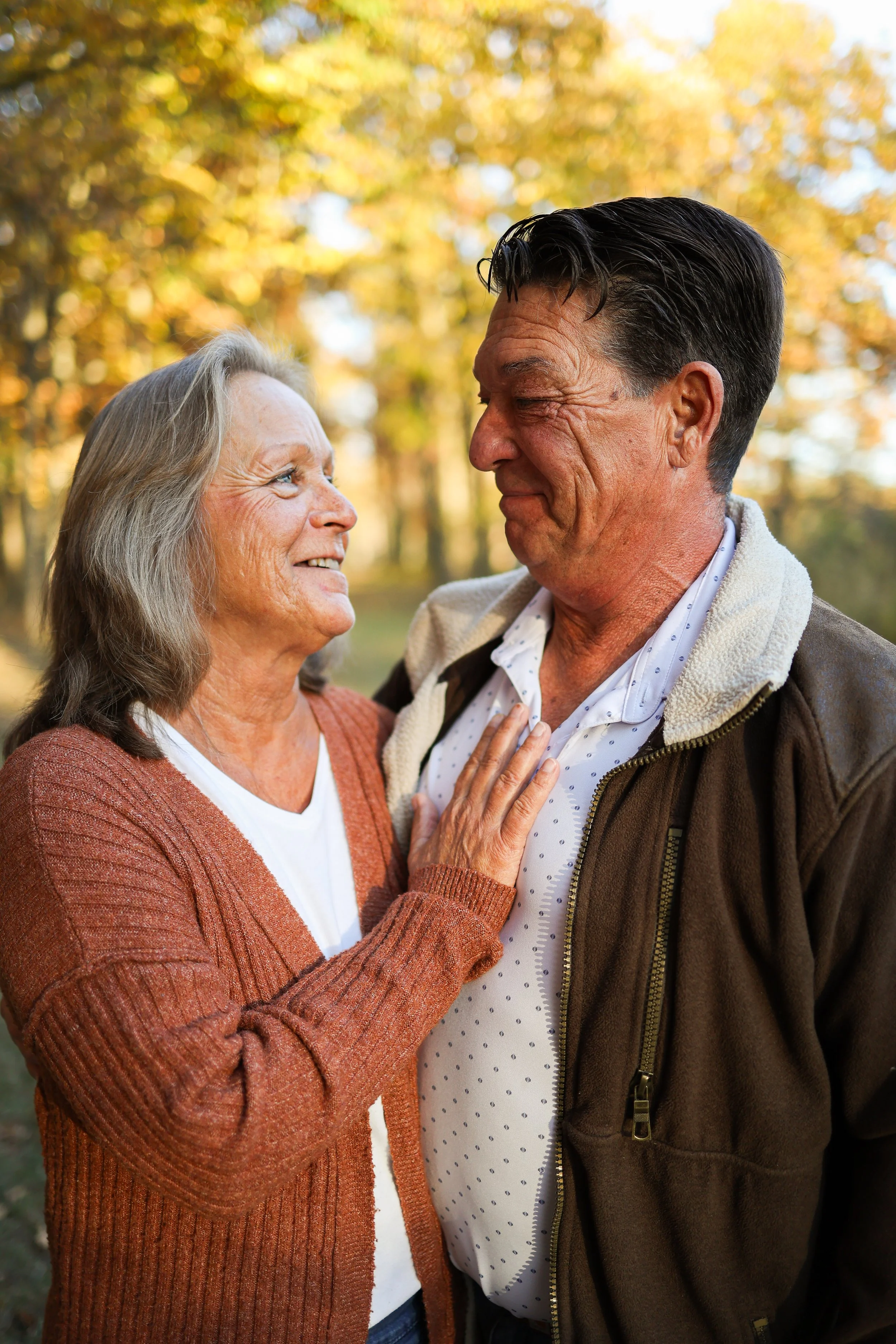 An elderly woman and man in an outdoor setting during autumn, smiling at each other, with fall foliage in the background.