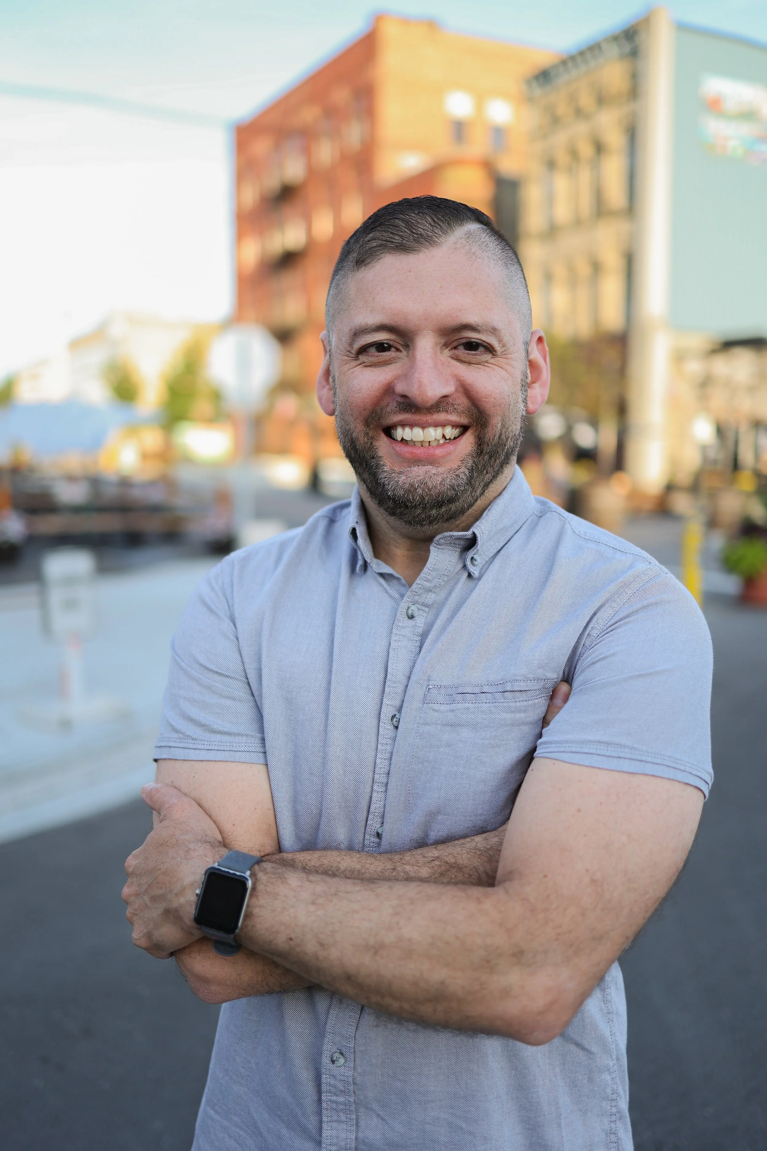 A man with short dark hair and a beard smiling in an outdoor urban setting with buildings in the background, wearing a light gray short-sleeve button-up shirt and a black smartwatch.