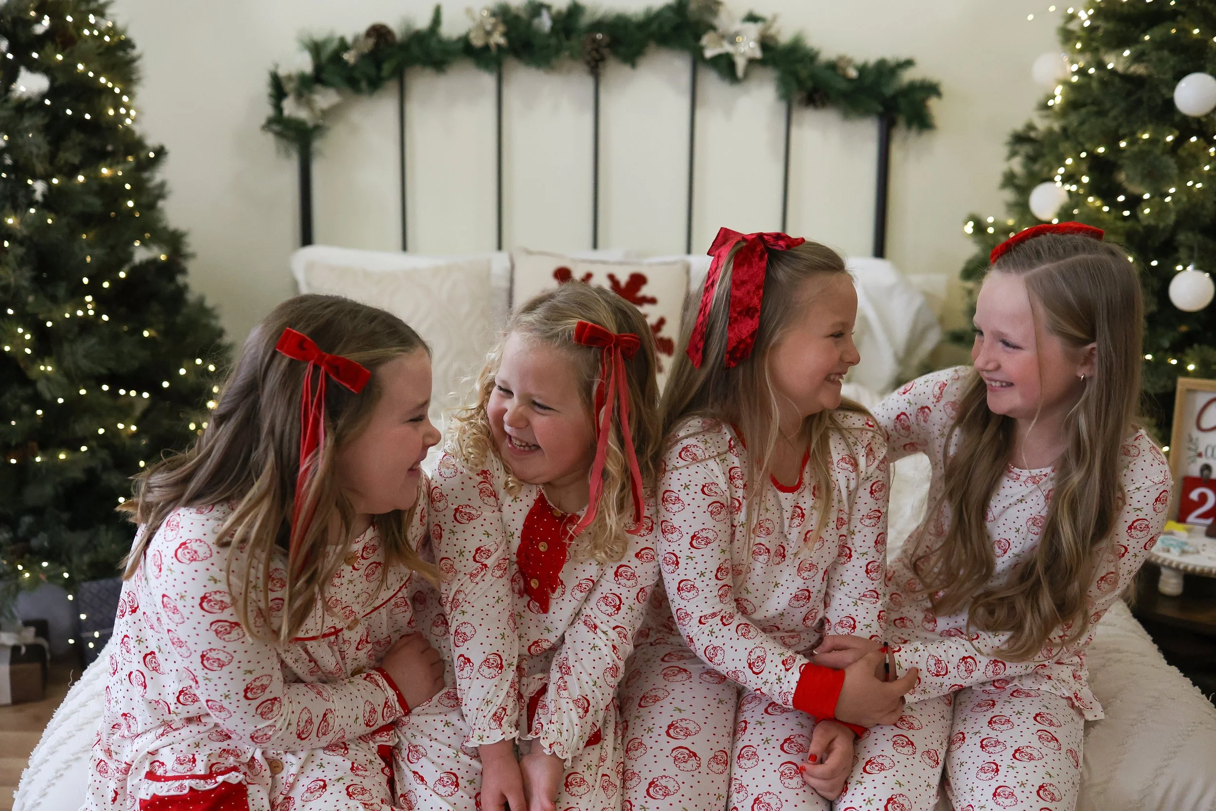 Four young girls wearing matching Christmas pajamas with red and white patterns sitting on a bed and laughing together in a decorated Christmas room.