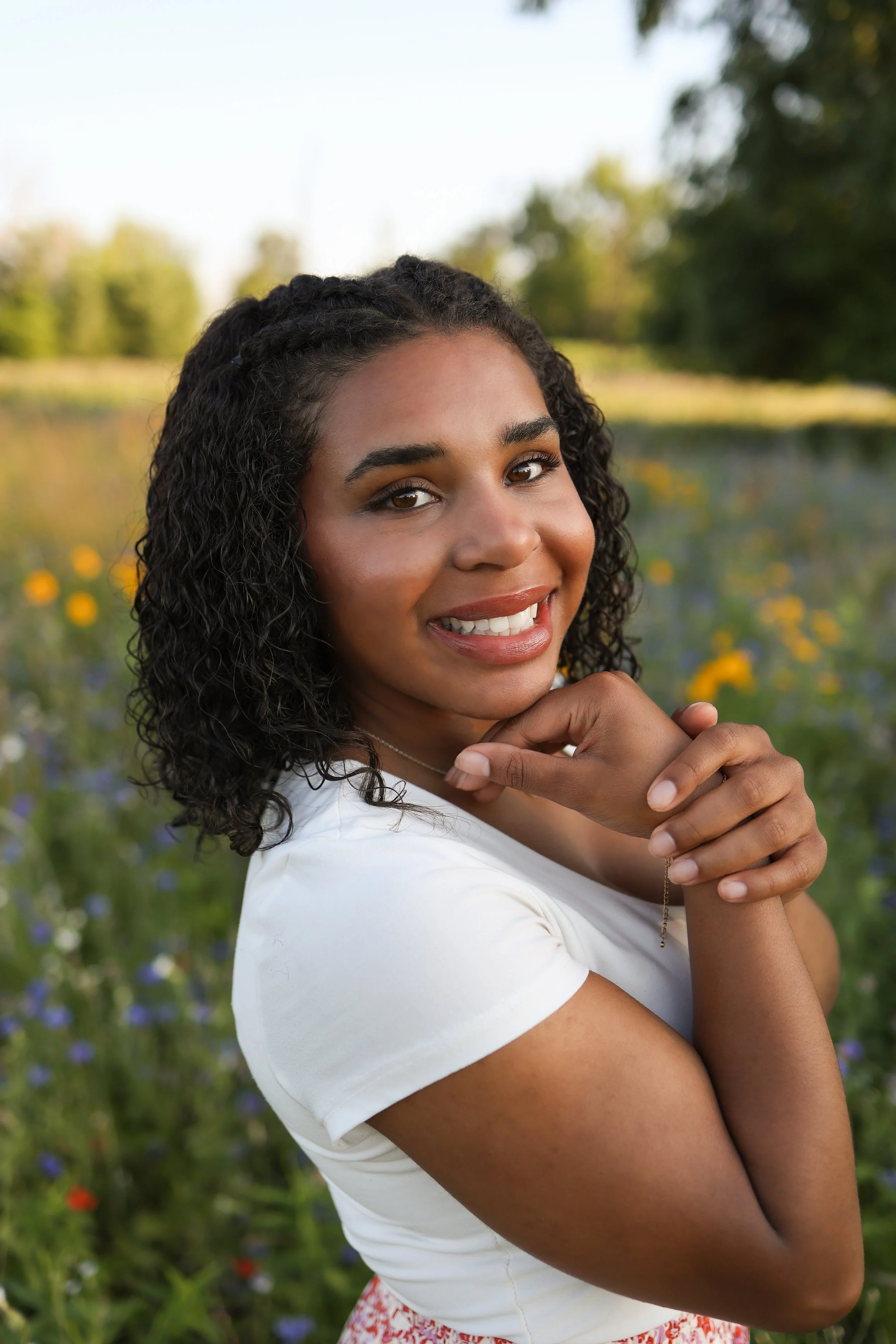 Young woman with curly dark hair smiling outdoors in a floral field.