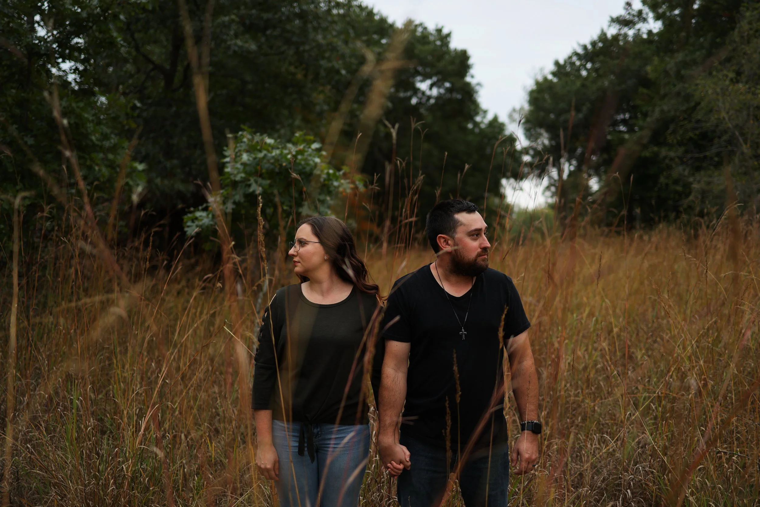 A couple holding hands and walking through tall grass in a forested area at dusk.