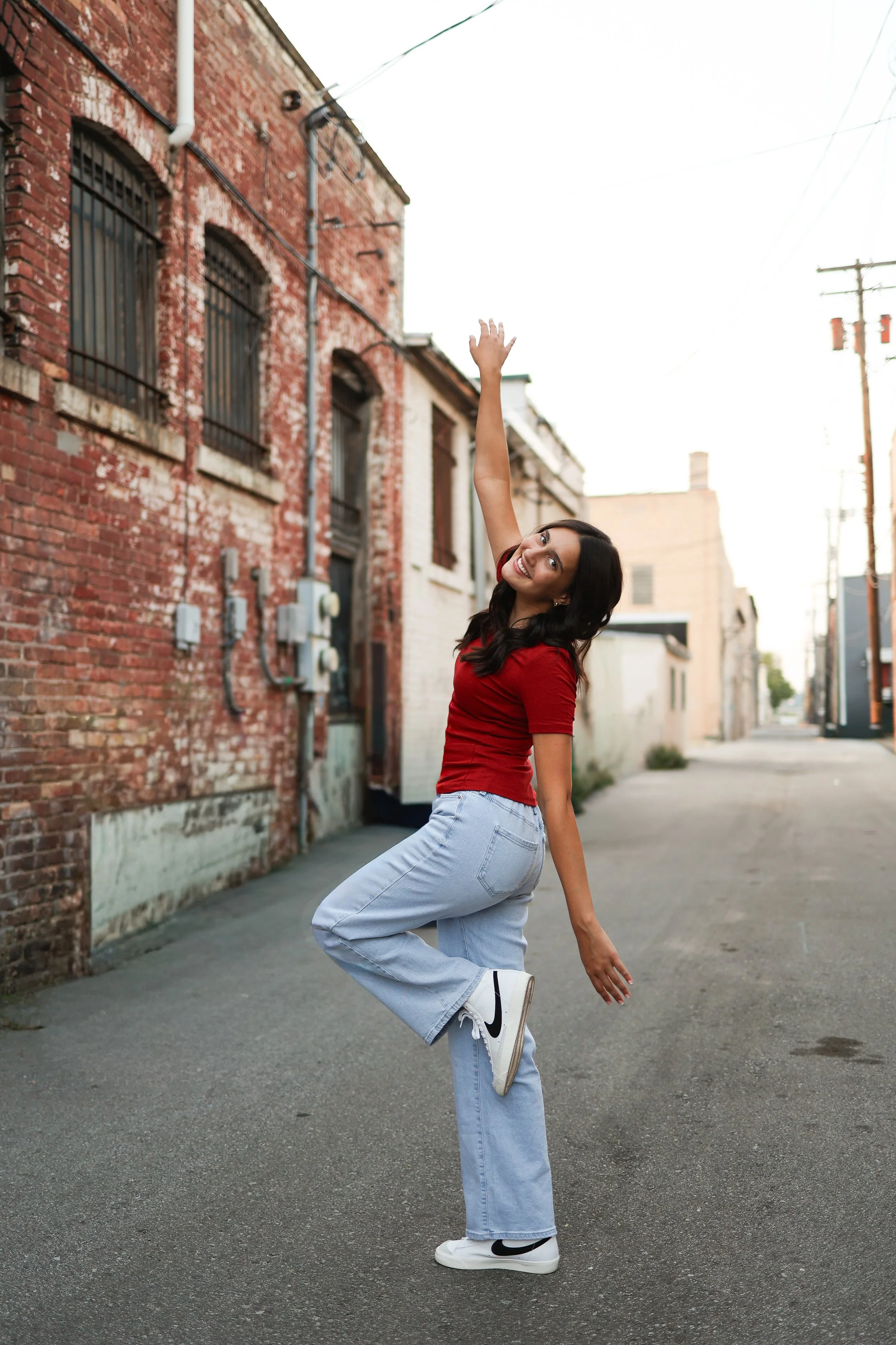 A young woman in a red shirt and light blue jeans is standing on an alleyway, smiling, with one arm raised and the other touching her foot, against a backdrop of old brick buildings.