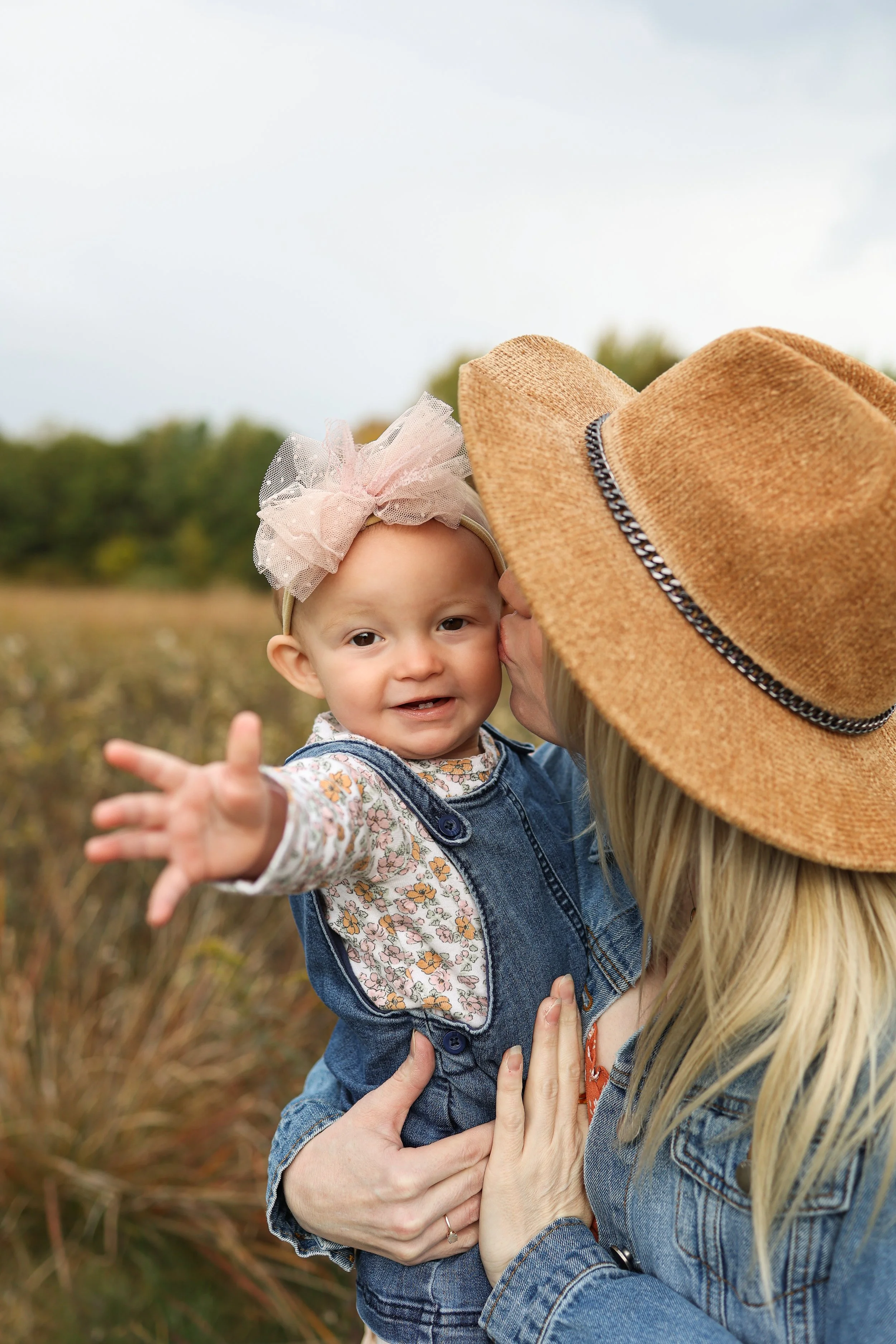 A woman holding a young girl outdoors in a field, with the woman wearing a wide-brimmed hat and the girl reaching out her hand toward the camera. The woman is whispering or kissing the girl on the cheek, and the girl is smiling.