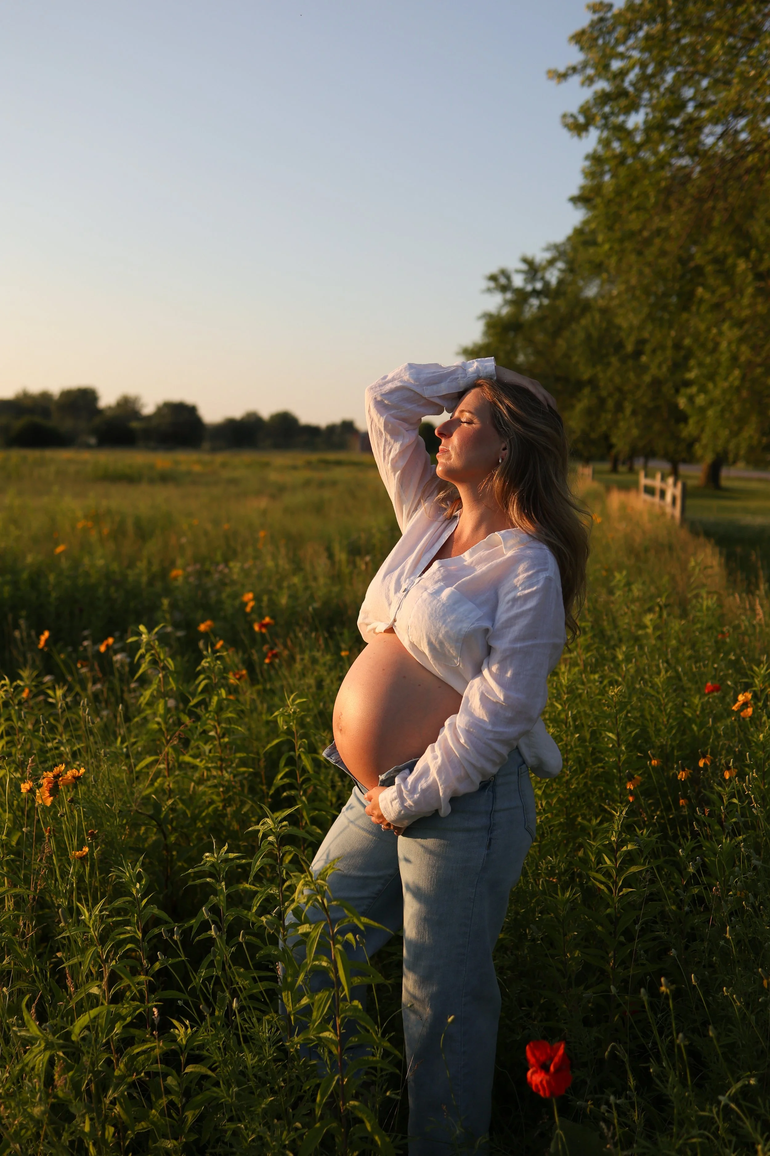 Pregnant woman standing in a field of flowers with the setting sun, wearing a white shirt and jeans, with one hand on her belly and the other resting on her head.