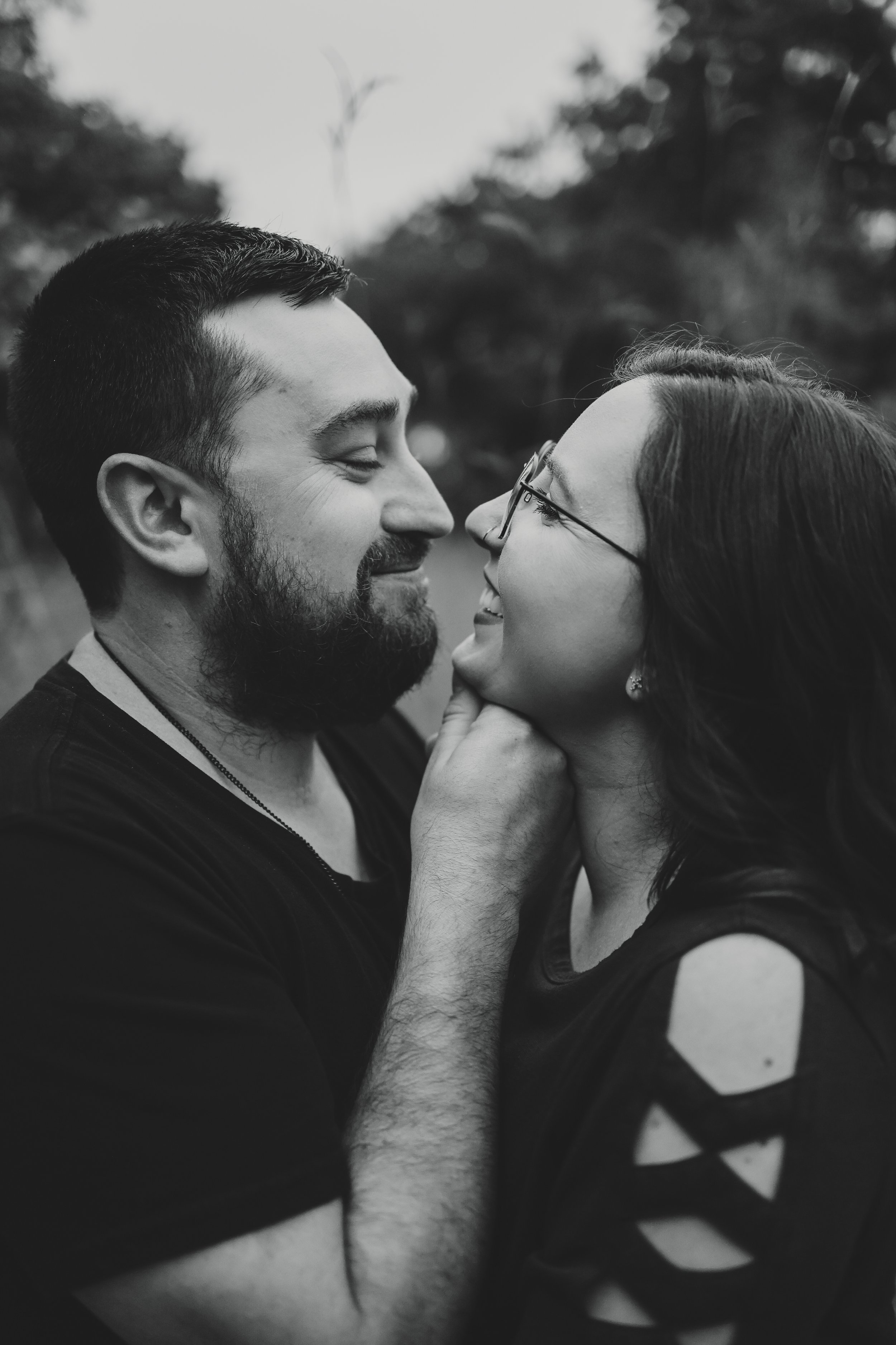 A black-and-white photo of a couple smiling close to each other outdoors, with the man holding the woman's chin, both appearing happy and affectionate.