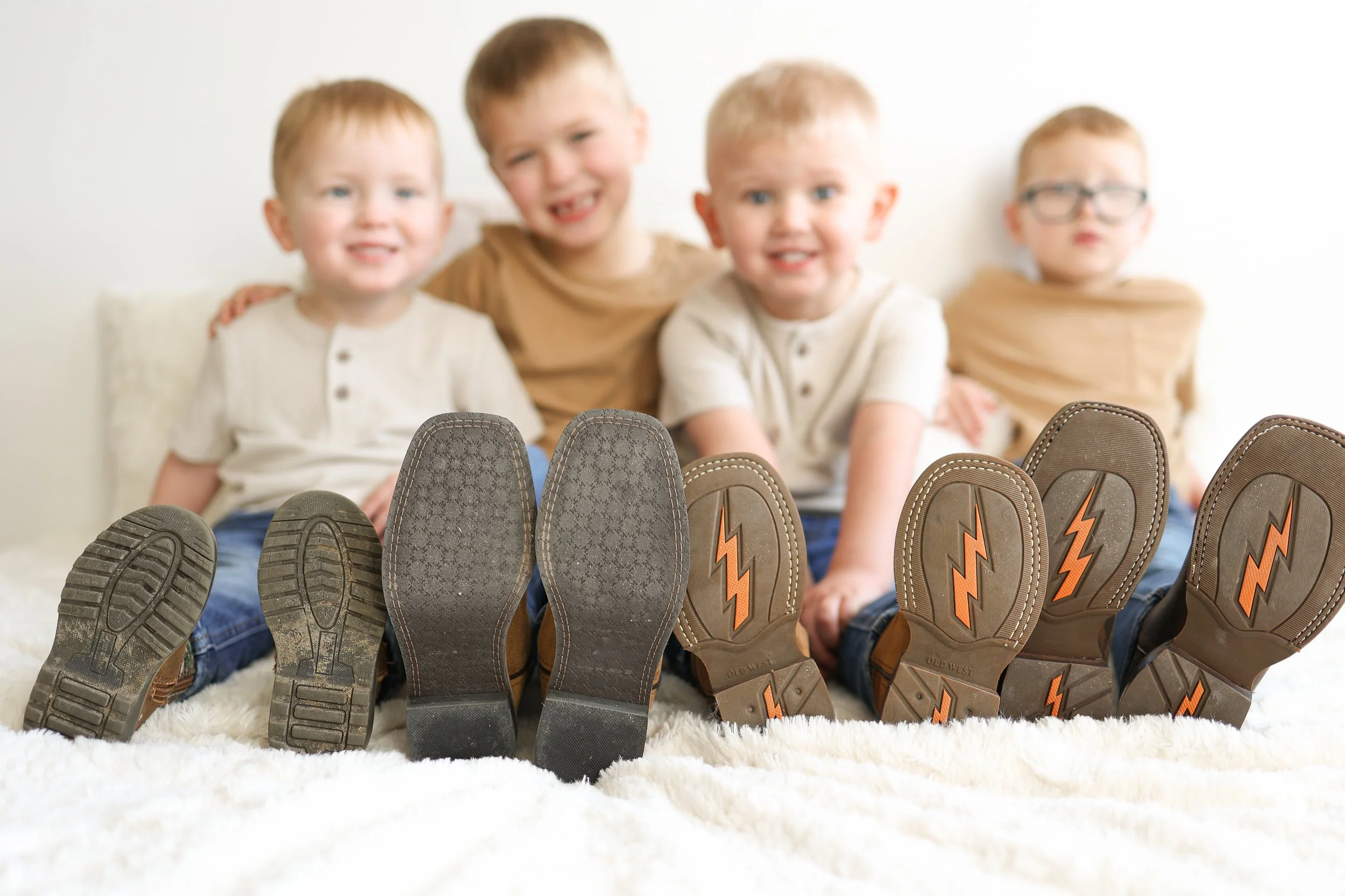 Four young boys sitting on a fluffy white blanket with their shoes and boots arranged in front of them, showing the soles and designs of their footwear.
