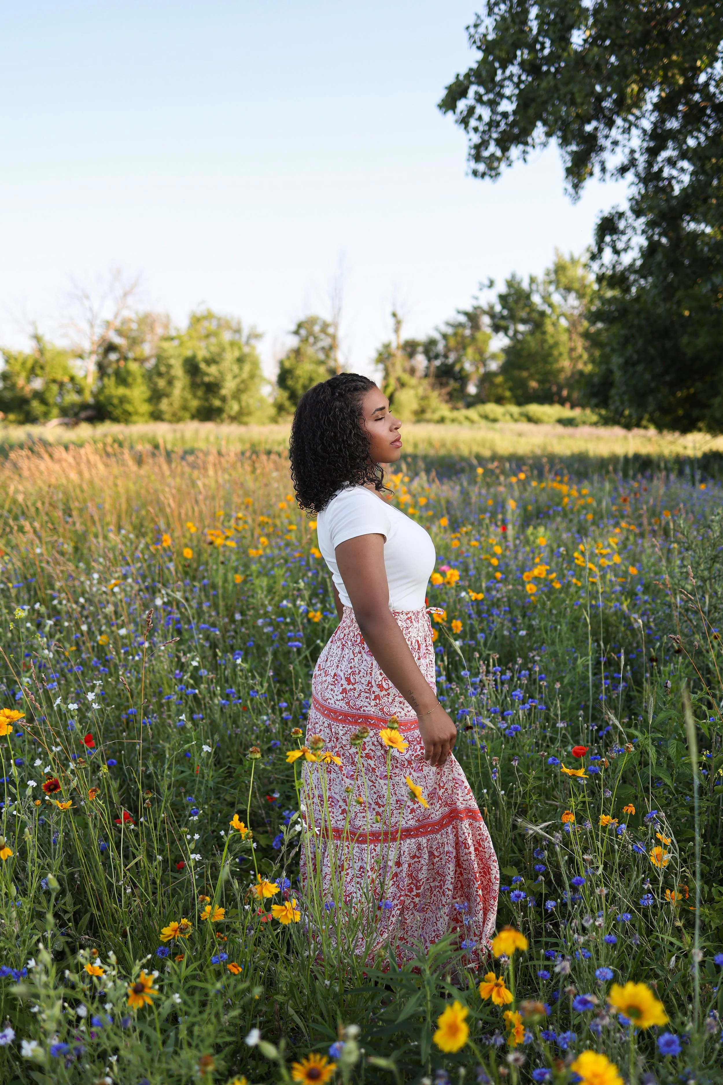 A woman standing in a field of wildflowers during daytime, with trees in the background and clear blue sky.