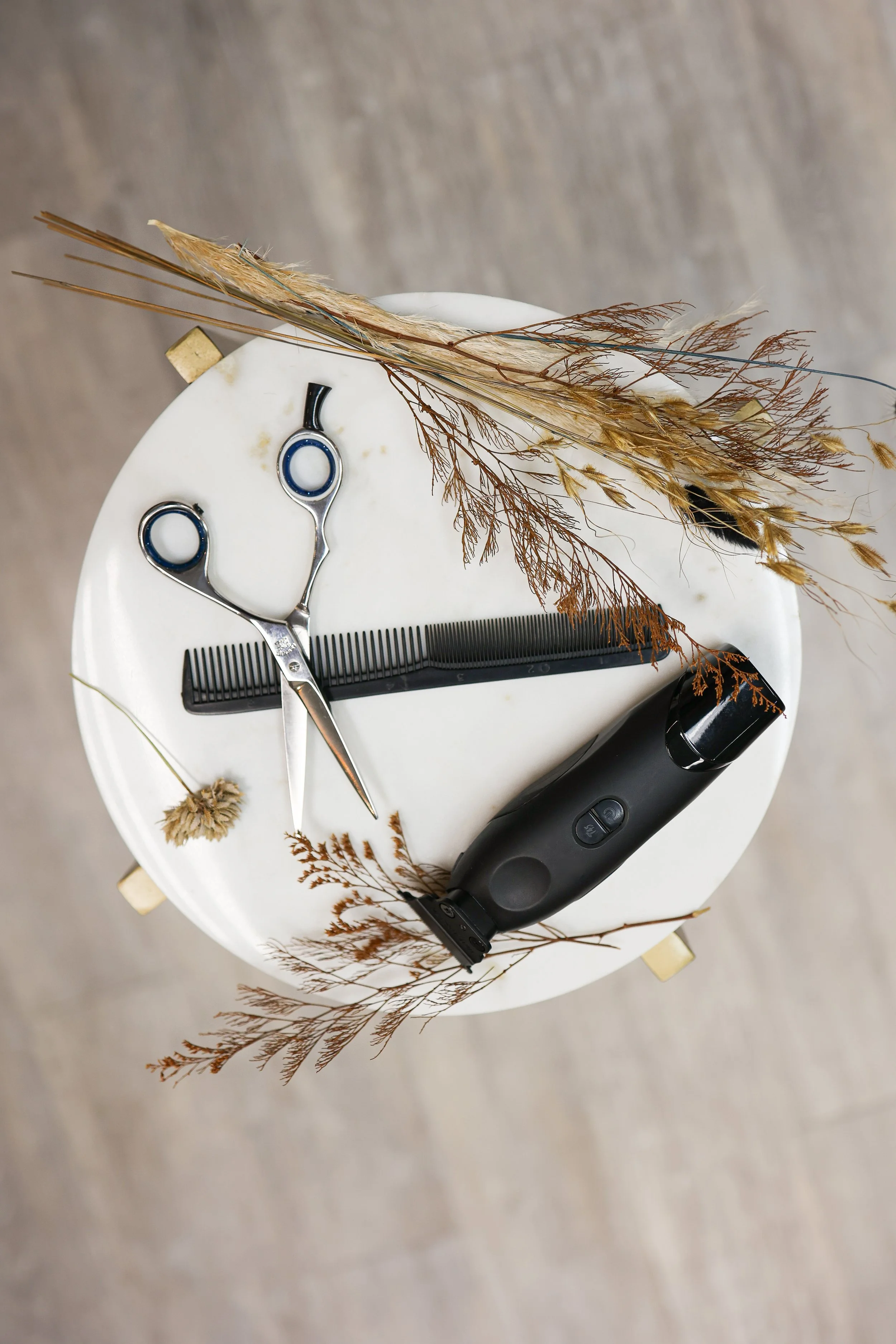 A round white table with hair cutting tools including a pair of scissors, a black comb, an electric hair trimmer, and dried decorative grasses arranged around the tools.