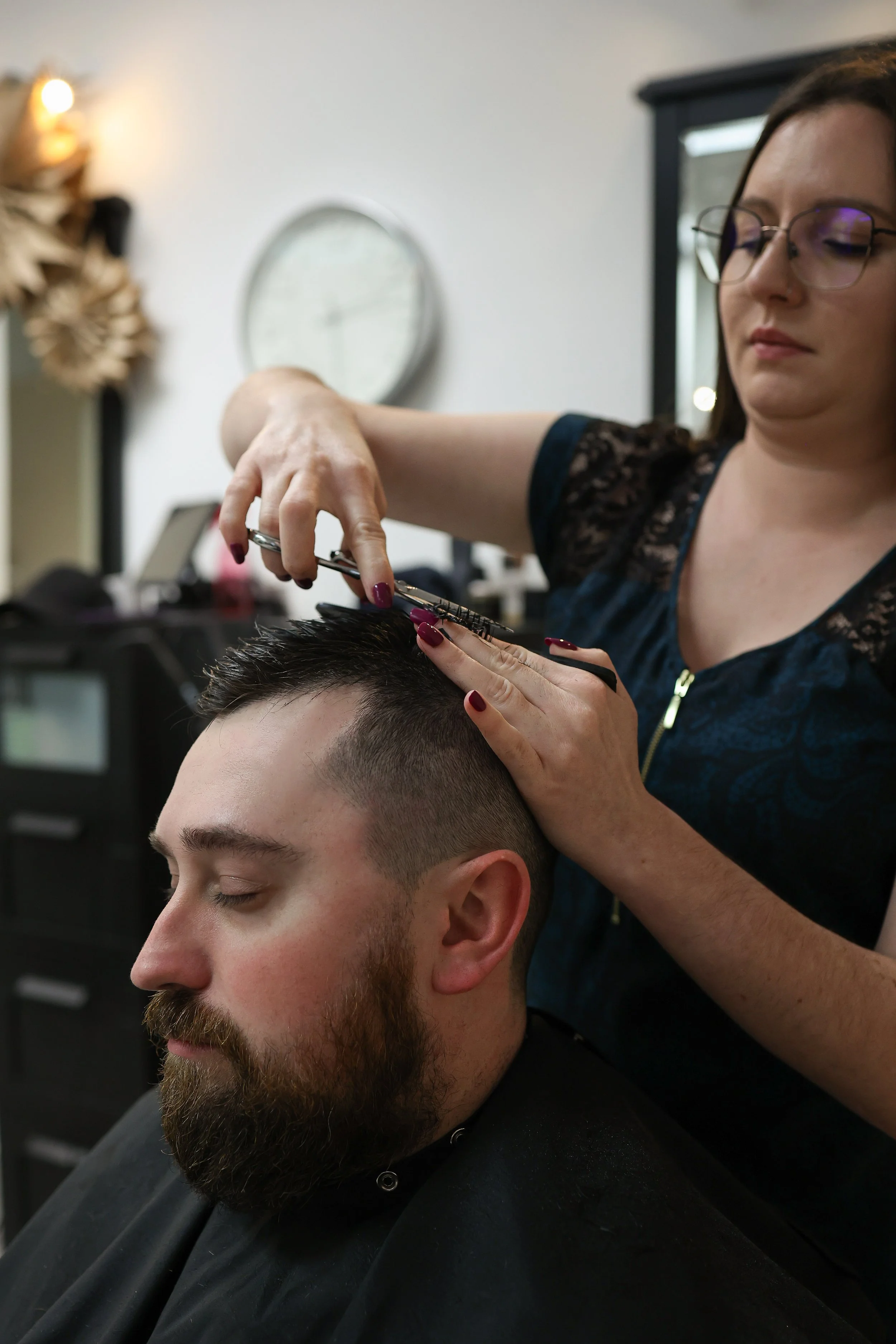 A woman with glasses and dark hair is giving a haircut to a man with a beard, who has his eyes closed and is seated in a salon chair.