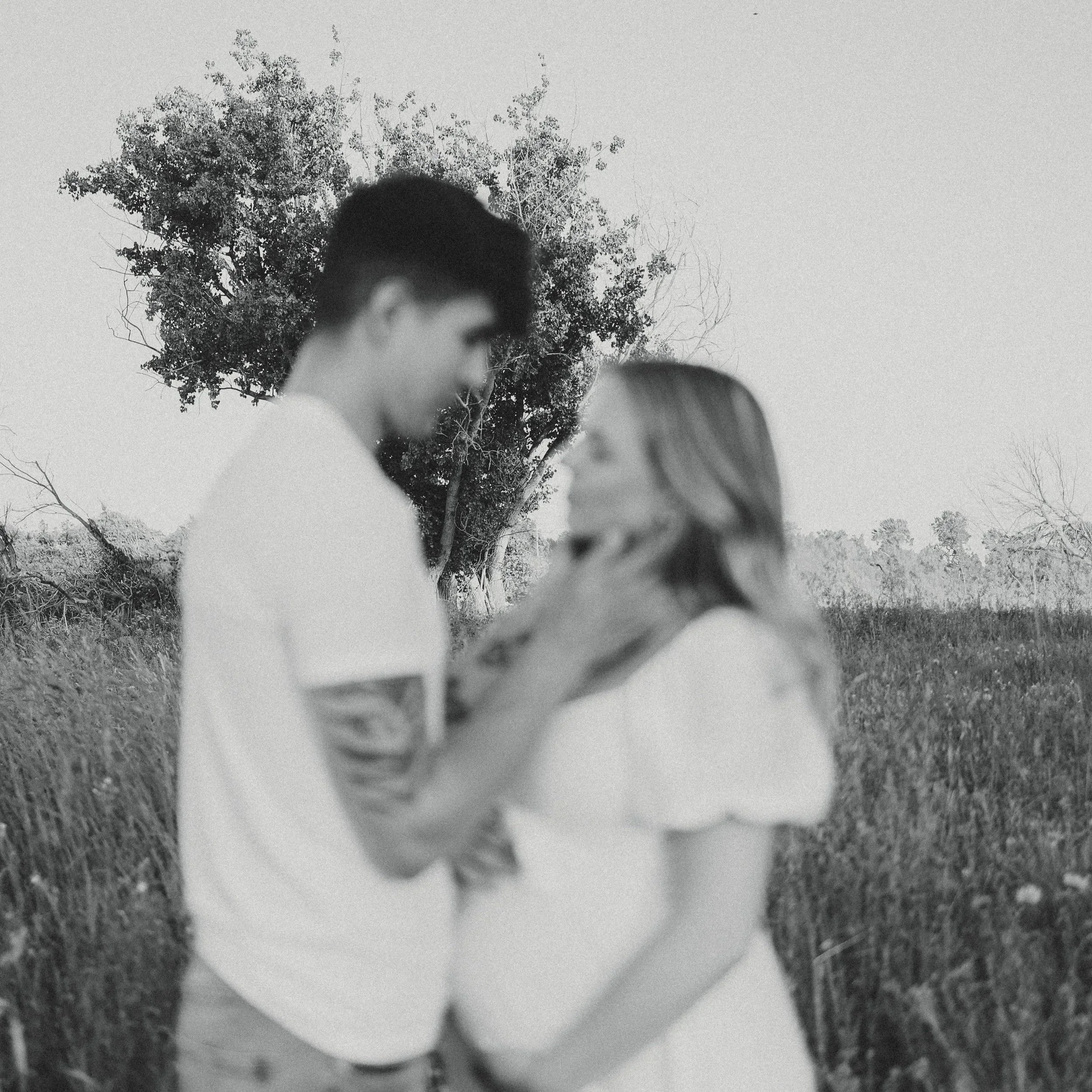 A black-and-white photo of a young couple standing close together in a field, with a large tree in the background. The man is touching the woman's chin gently, and they are looking at each other.