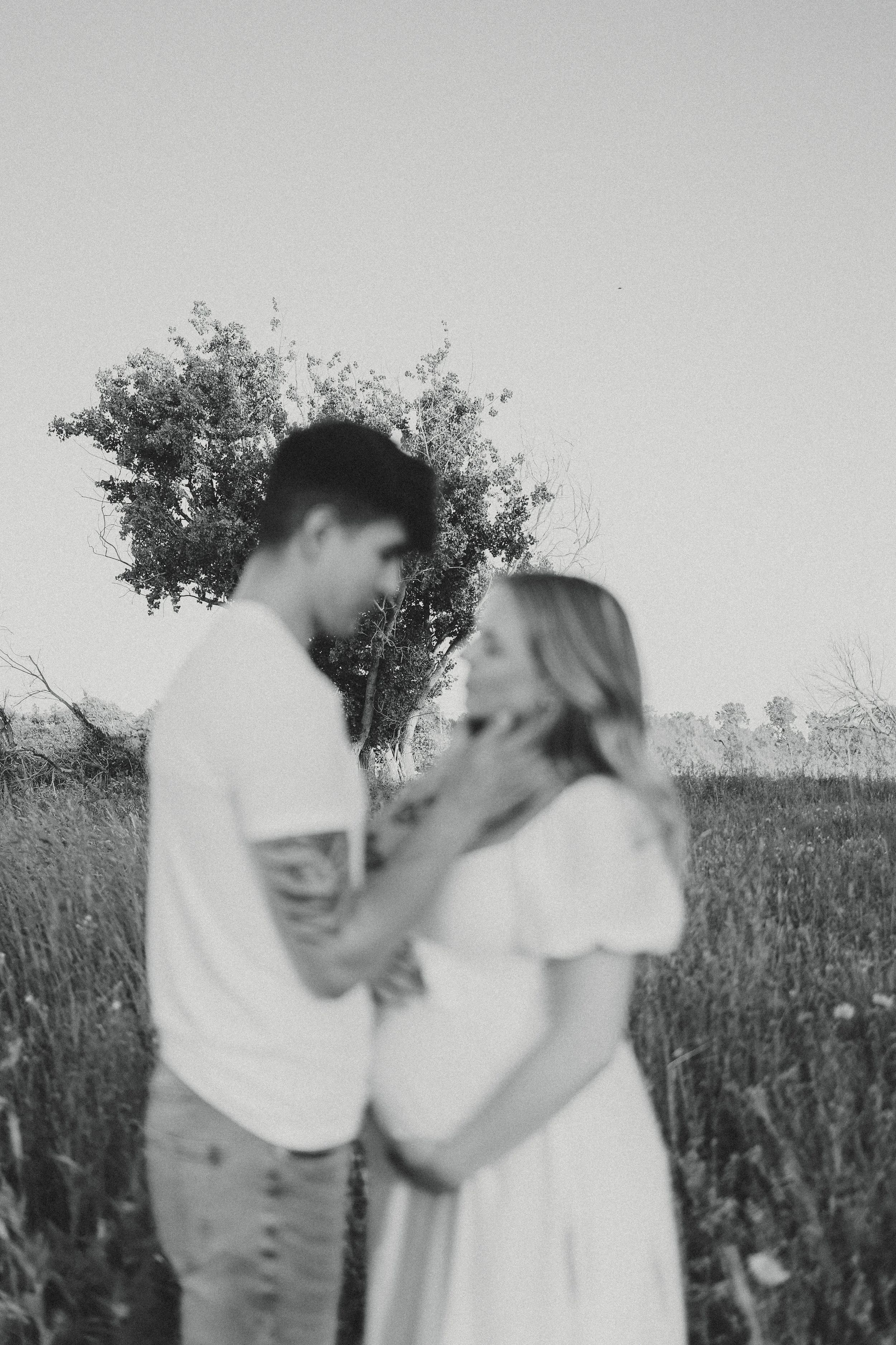 A black and white photo of a couple standing close together outdoors, with the man touching the woman's face, in a field with trees in the background.