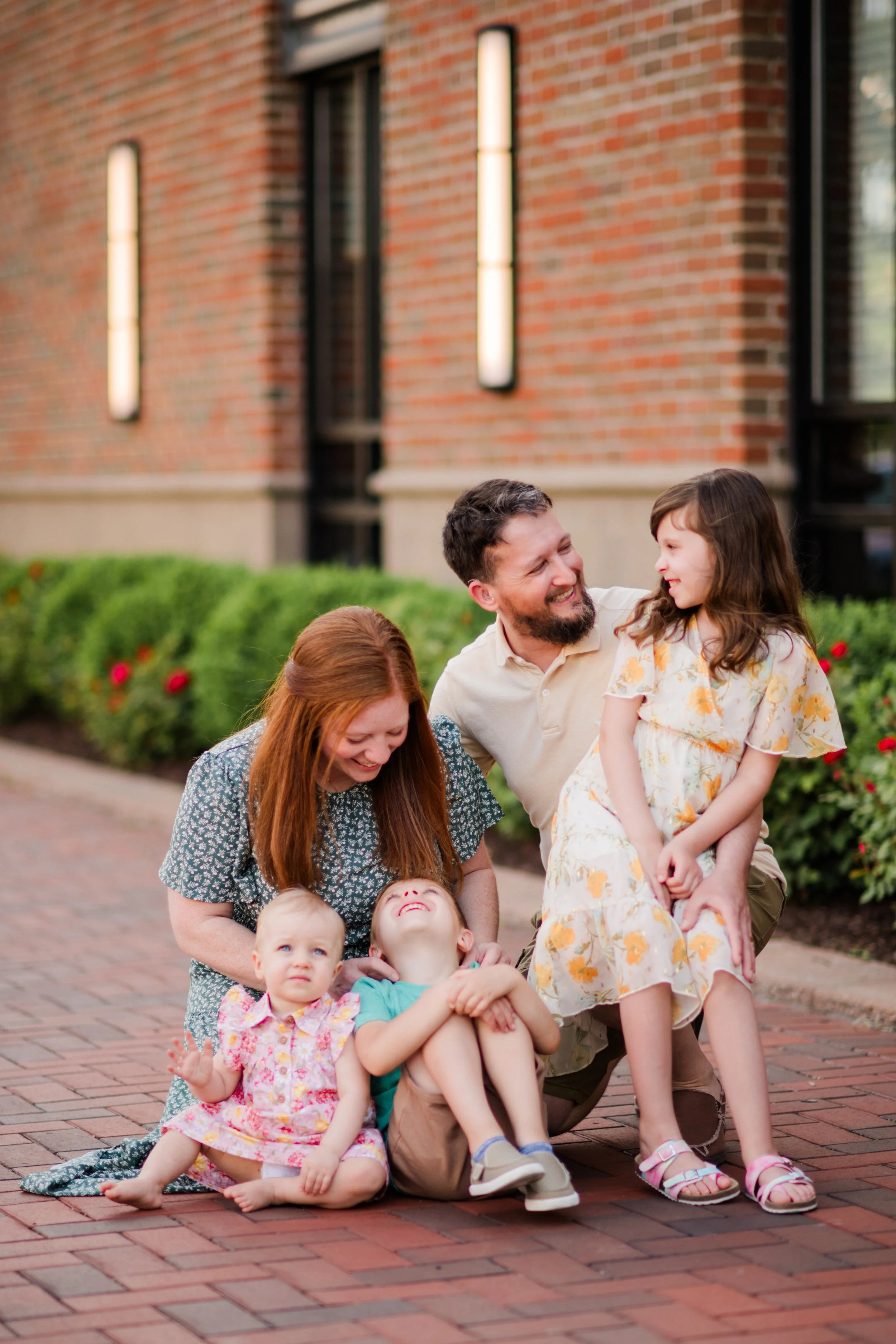A happy family of five sitting on a brick sidewalk in front of a brick building with greenery.