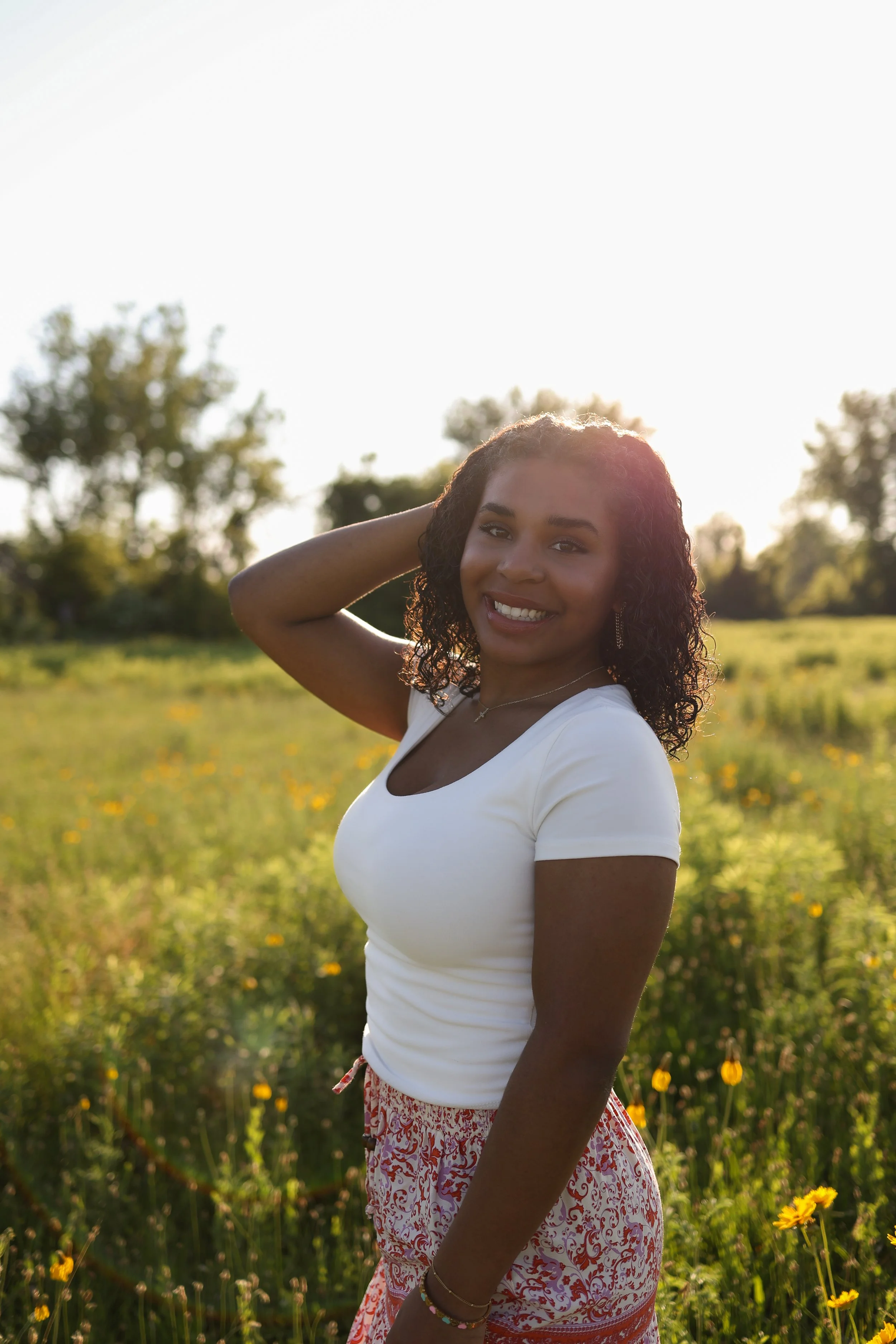 A young African America woman with curly hair smiling and posing outdoors in a field of yellow flowers during sunset for a senior session.