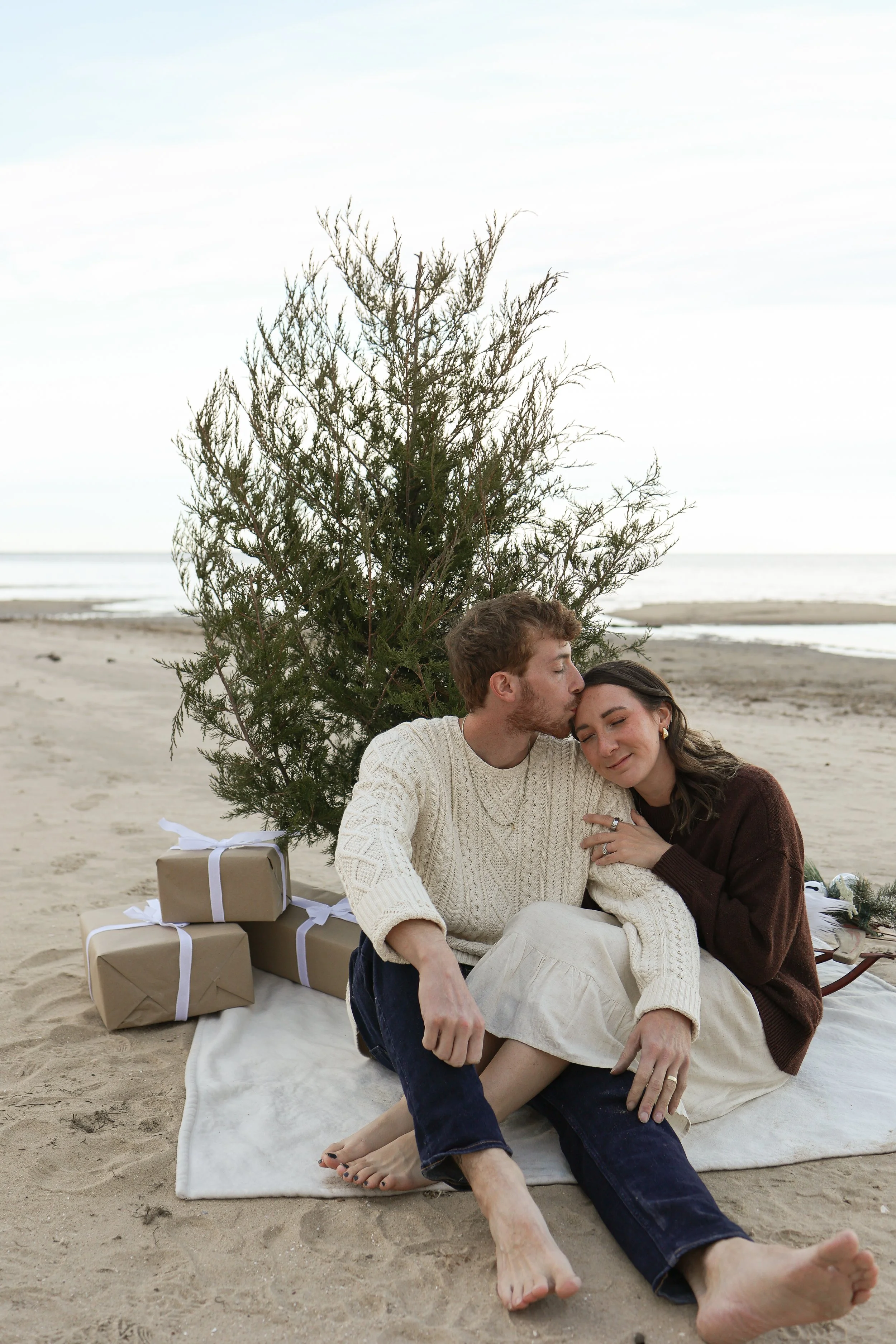 A couple sitting on a white blanket on the beach, surrounded by wrapped Christmas presents, with a small Christmas tree behind them. The man is kissing the woman's forehead while she smiles with her eyes closed.