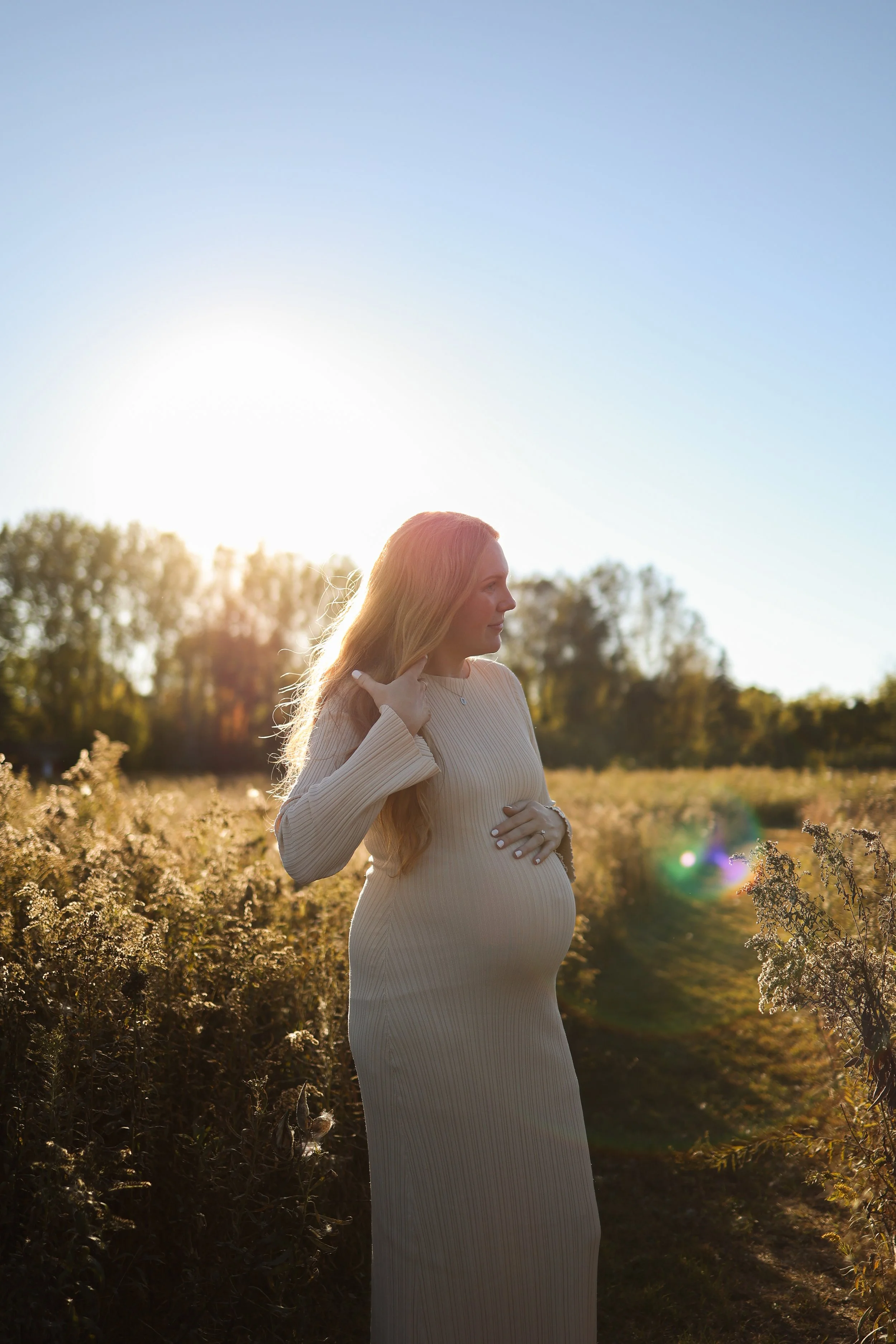 Pregnant woman with long hair standing in a field at sunset, wearing a light-colored, long-sleeved dress, touching her belly and looking to the side.