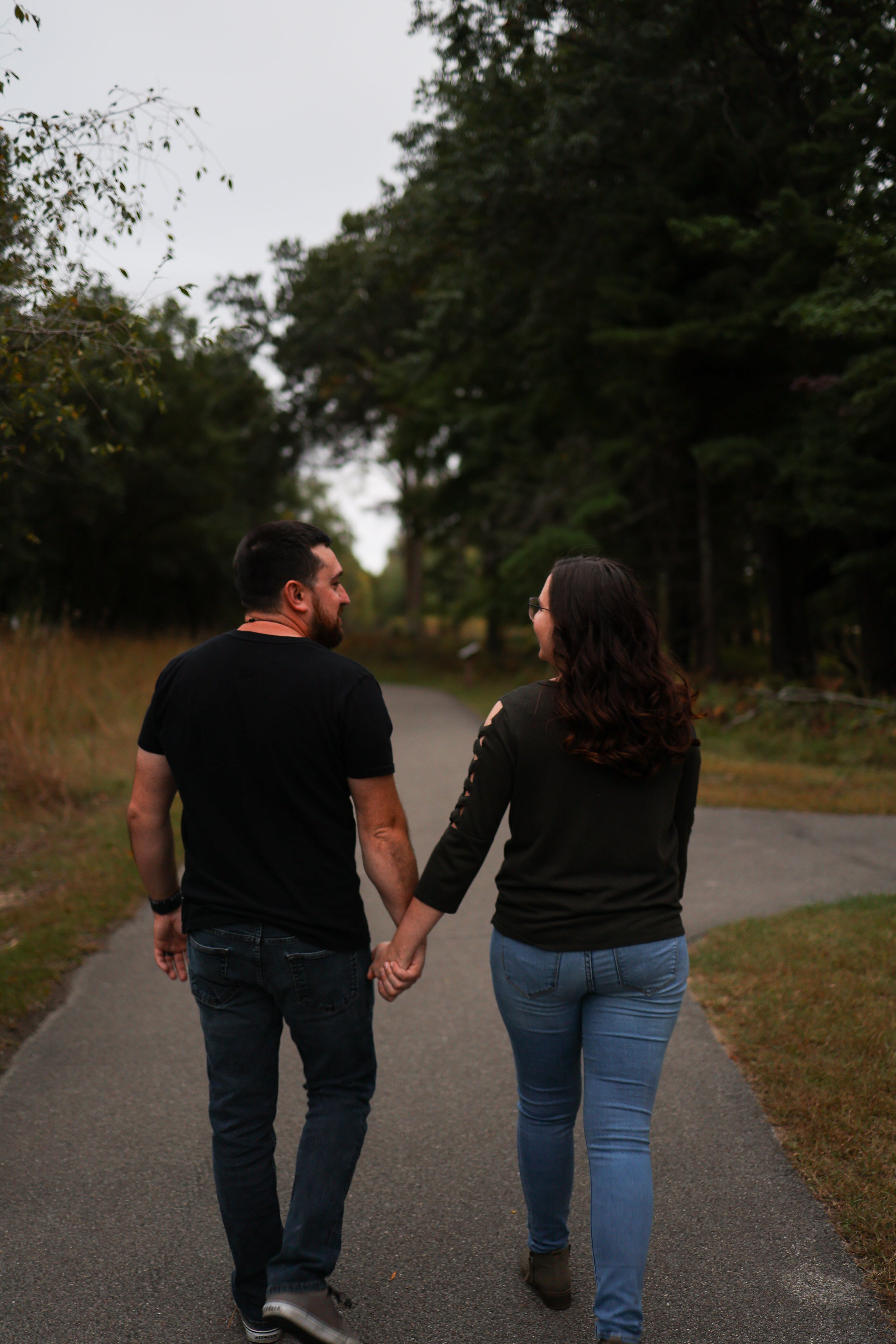 A couple holding hands and walking on a paved path through a park or wooded area with trees.
