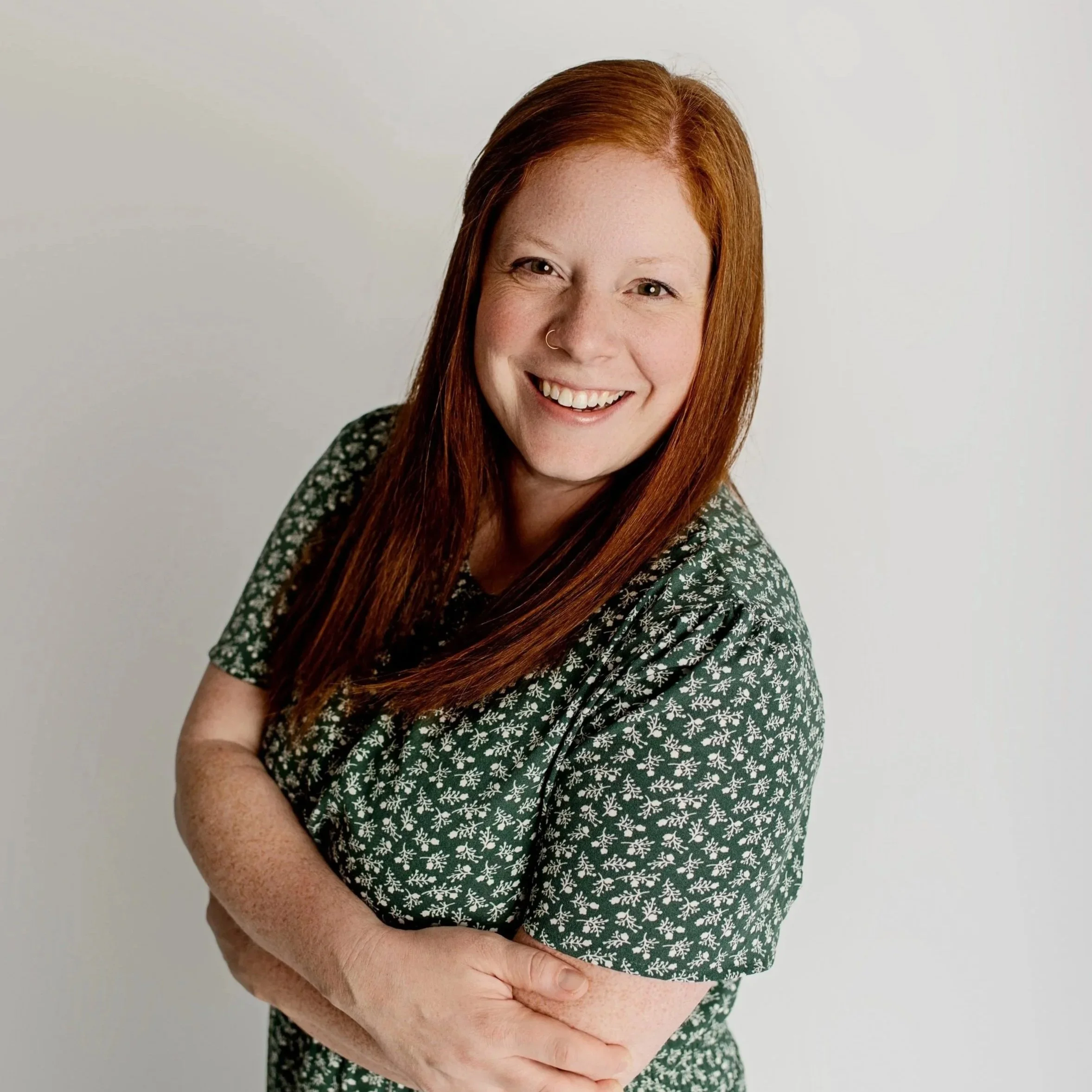 A woman with red hair smiling and wearing a green patterned shirt, standing against a plain white background.