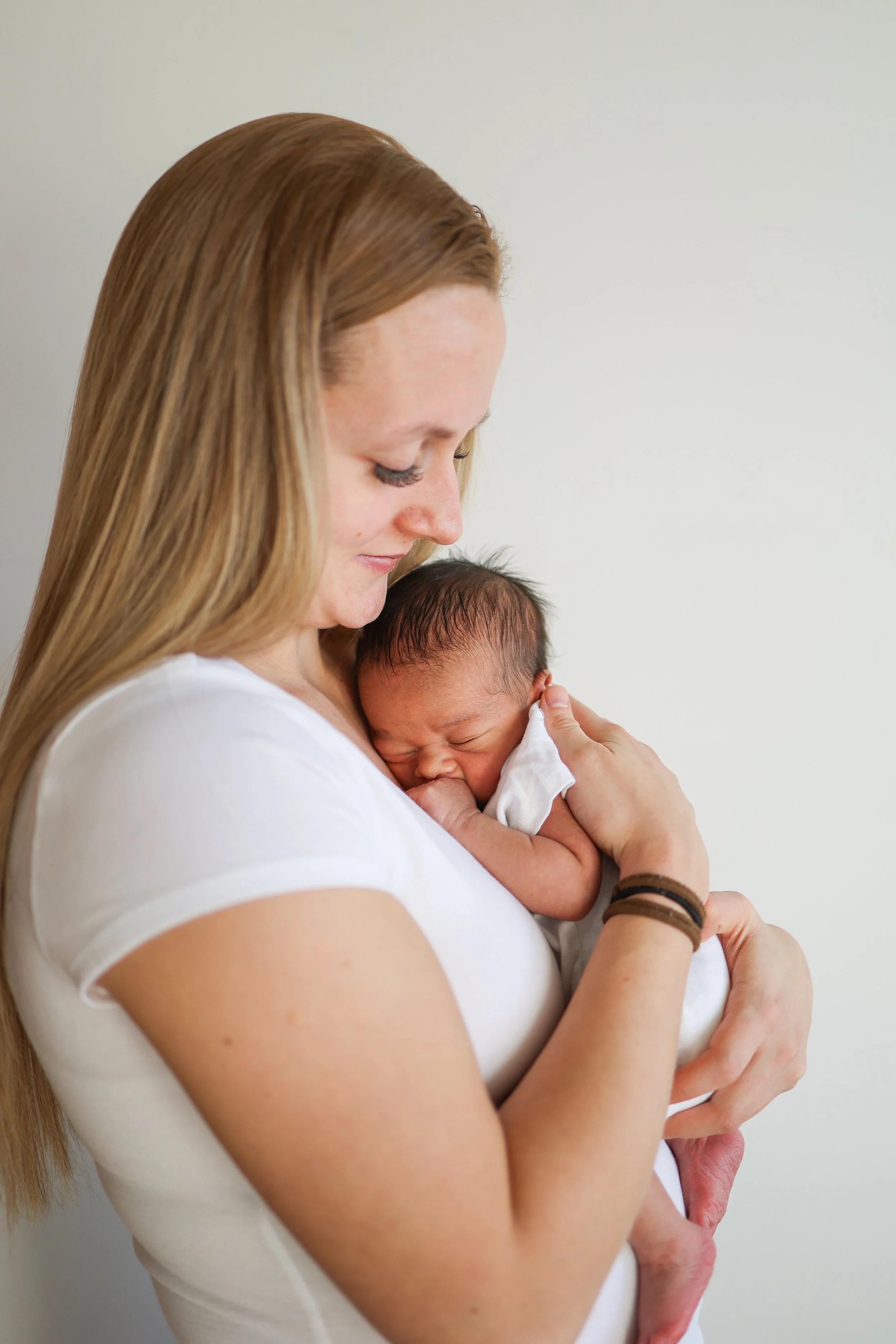 A woman with long red hair holding a newborn baby close to her chest, with the baby resting its head on her shoulder, in front of a plain, light-colored wall.