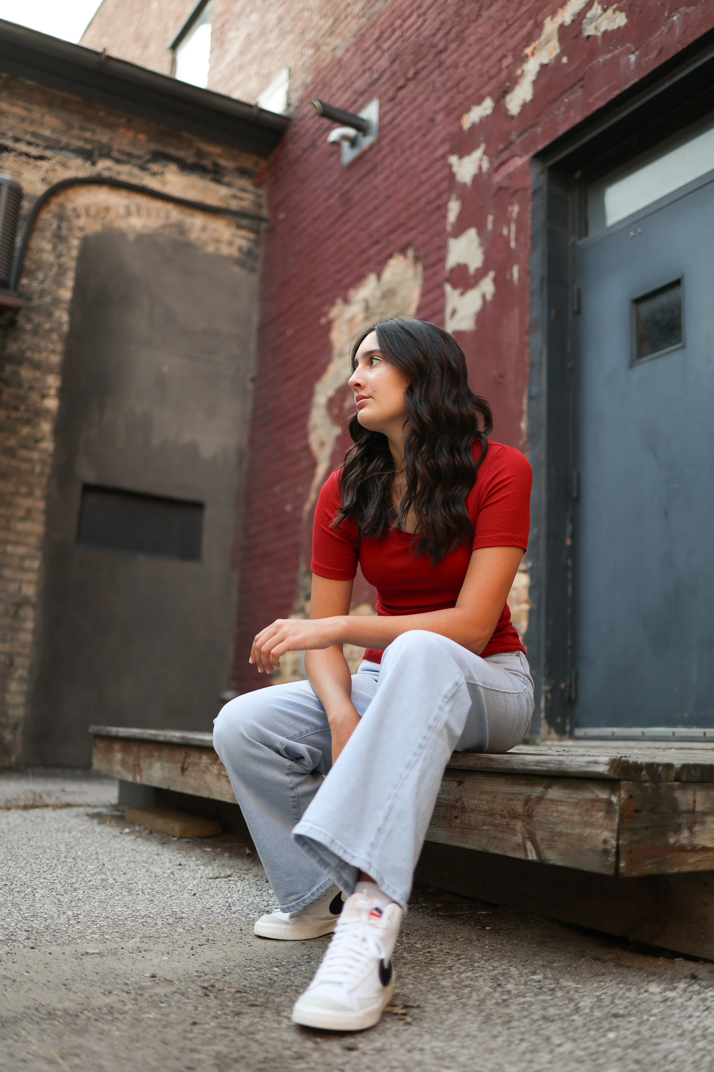 Young woman with dark wavy hair wearing a red shirt, light gray jeans, and white sneakers sitting on a wooden platform in an alleyway with weathered brick walls and a gray metal door.