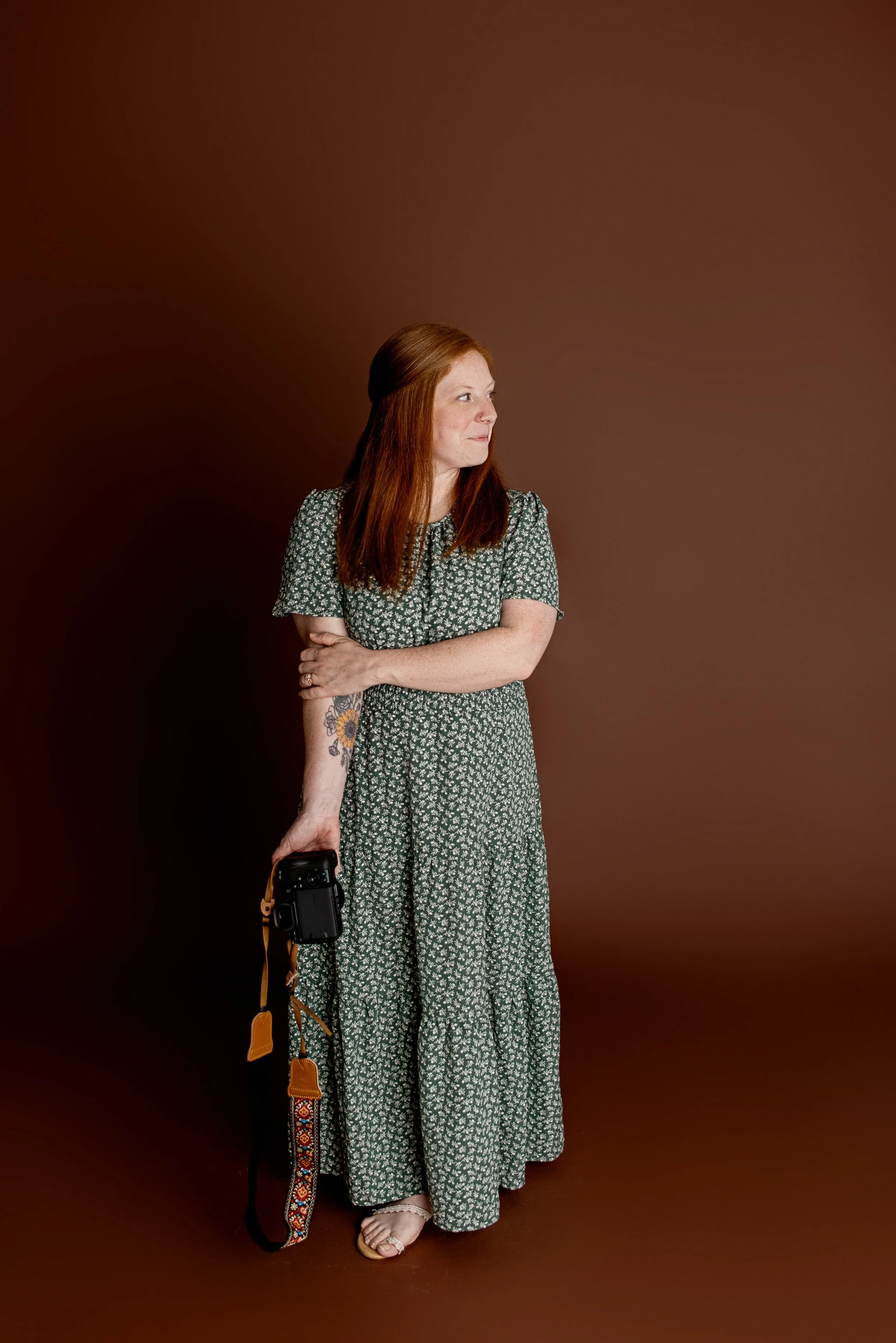 A woman with long red hair, wearing a green floral dress, holding a camera with a patterned strap, standing against a brown background.