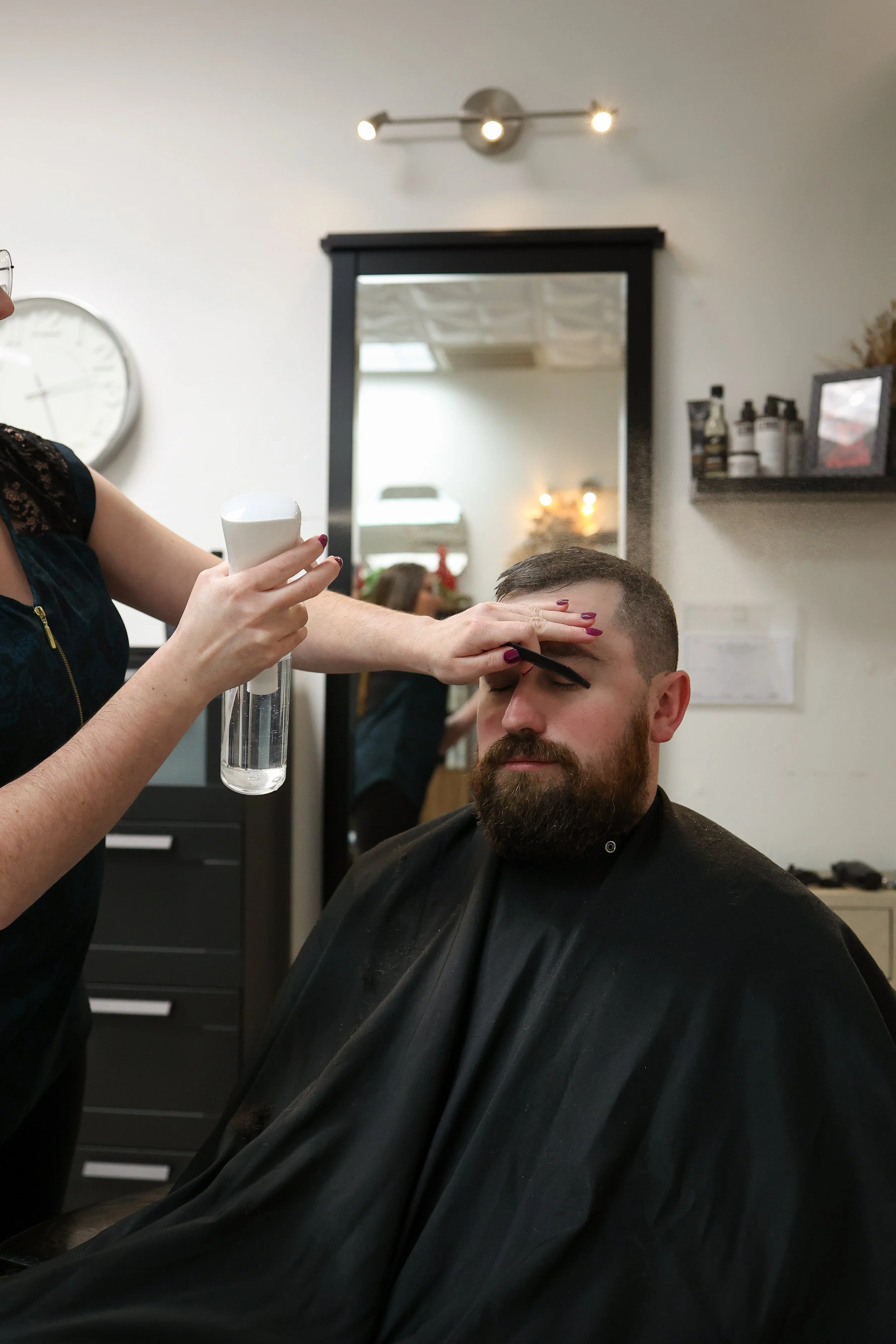A man with a beard and a black cape getting the full treatment at a salon or barbershop, while a stylist sprays water on his hair.