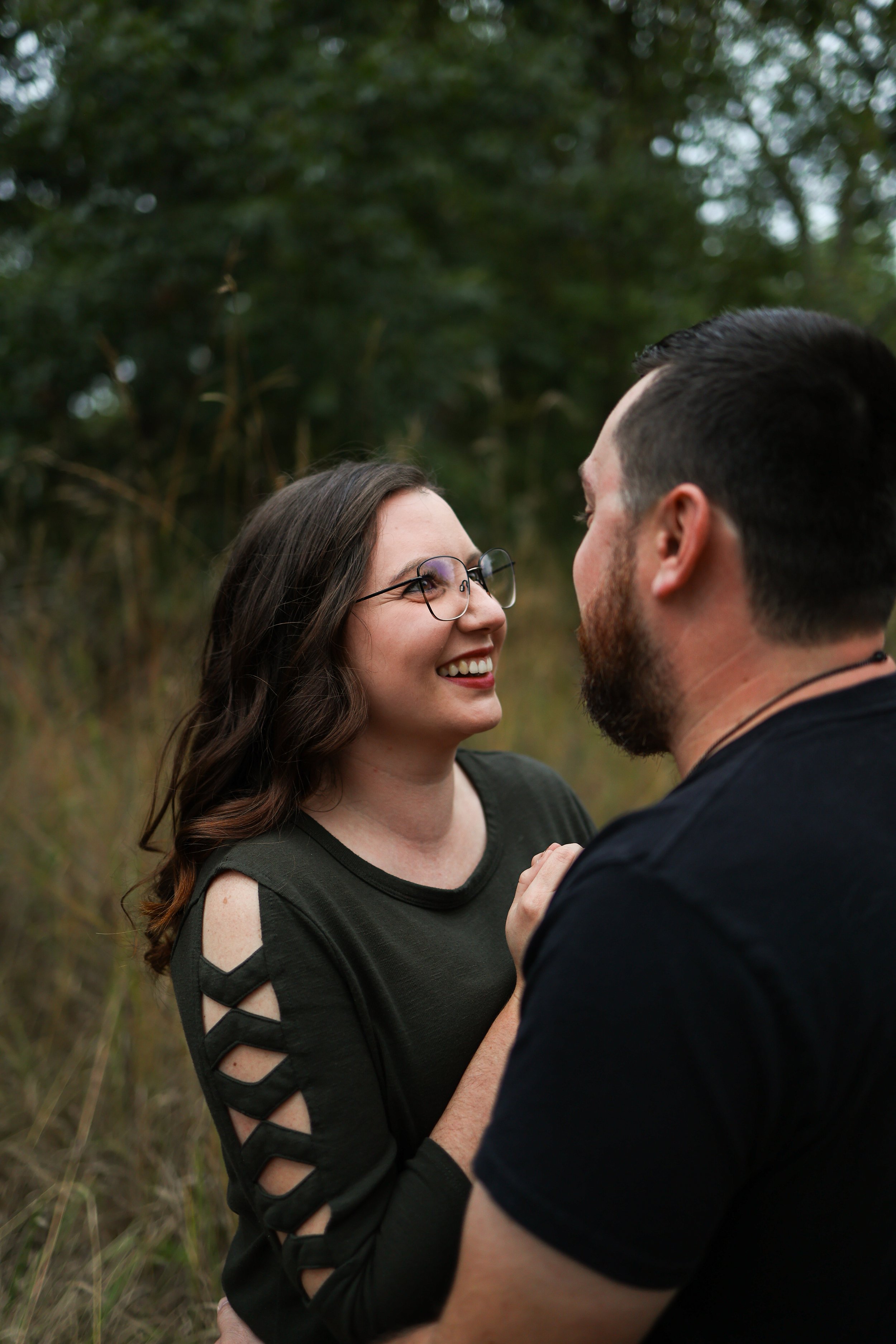 A couple standing close together outdoors in a wooded area, smiling at each other.